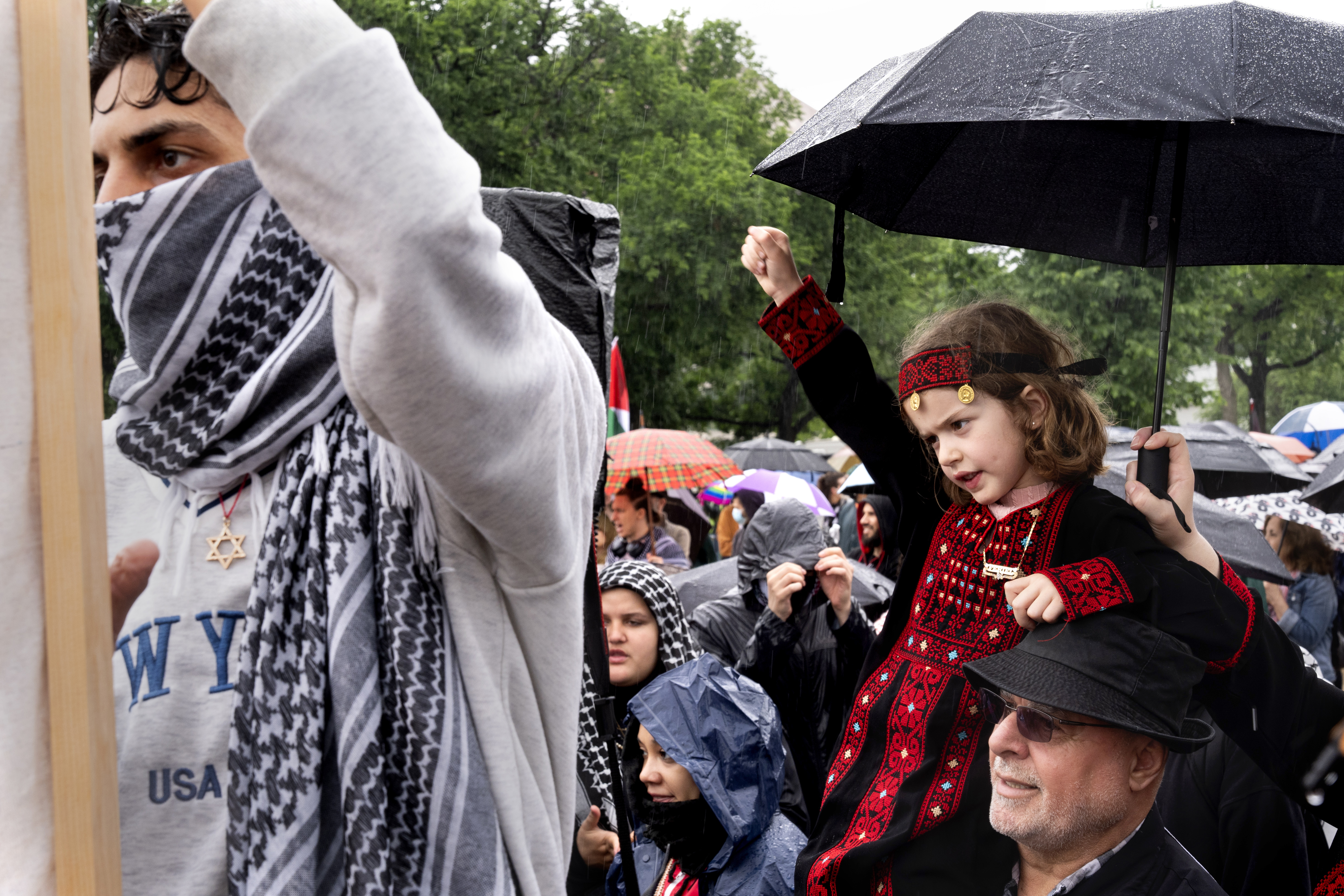 A young man wearing a keffiyeh and a Jewish star necklace, left, holds a banner next to a 7-year-old from Springfield, Va., wearing a traditional outfit on the shoulders of her grandfather, who is Palestinian, during a pro-Palestinian rally, Saturday, May 18, 2024, on the National Mall near the Capitol in Washington. (AP Photo/Jacquelyn Martin)