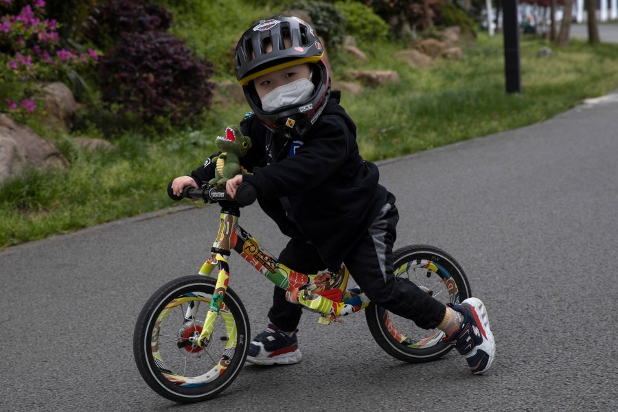 FILE - A child wearing a mask pushes his bike during a quiet day at the East Lake Park in Wuhan in central China's Hubei province, April 2, 2020. Miniature bikes with no pedals seem to be everywhere these days, and the kids riding them are getting younger and younger. Experts recommend starting with those so-called balance bikes at a younger-than-expected age, possibly even less than a year old. (AP Photo/Ng Han Guan, File)