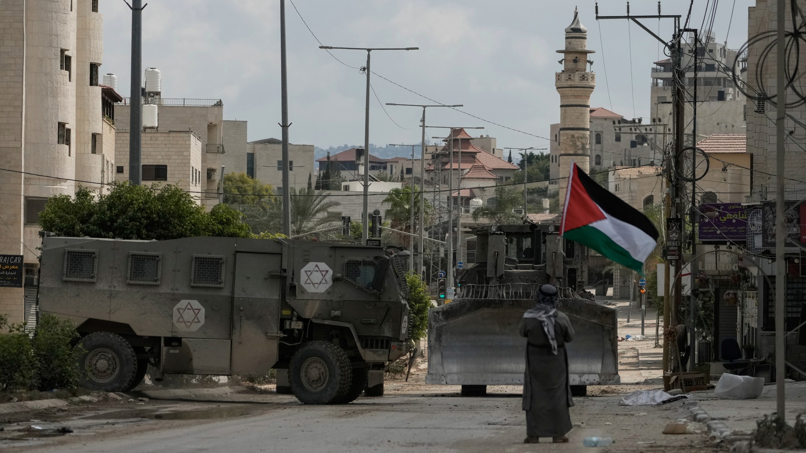 Palestinian activist Khairi Hanoon waves the Palestinian flag as a convoy of Israeli military armored vehicles drives by during an army raid in Tulkarem, West Bank, on Tuesday, Sept. 3, 2024. (AP Photo/Majdi Mohammed)