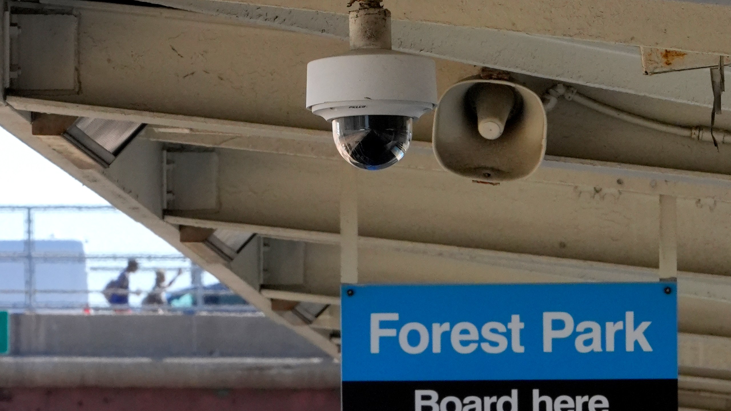 A security camera and speaker hang from the ceiling of the Chicago Transit Authority Harlem Ave. station over the rails that head West to Forest Park, Ill., station as two pedestrians walk toward the station, Tuesday, Sept. 3, 2024, in Forest Park, Ill. (AP Photo/Charles Rex Arbogast)