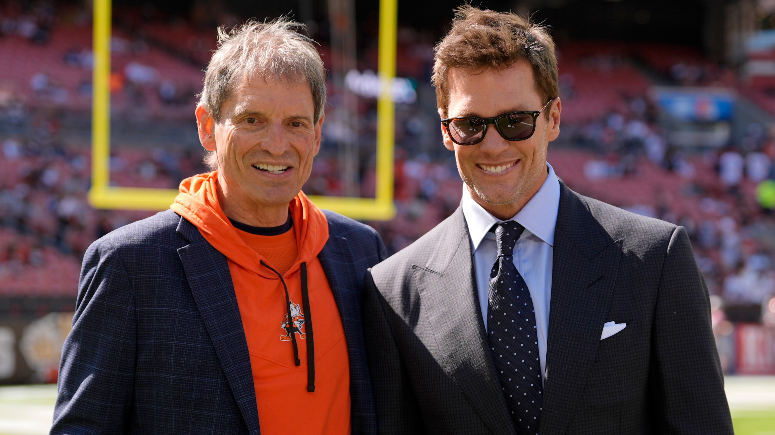 Former NFL players Bernie Kosar, left, and Tom Brady, right, pose for a photo as they talk on the field during warmups before a football game between the Dallas Cowboys and the Cleveland Browns in Cleveland, Sunday, Sept. 8, 2024. (AP Photo/Sue Ogrocki)