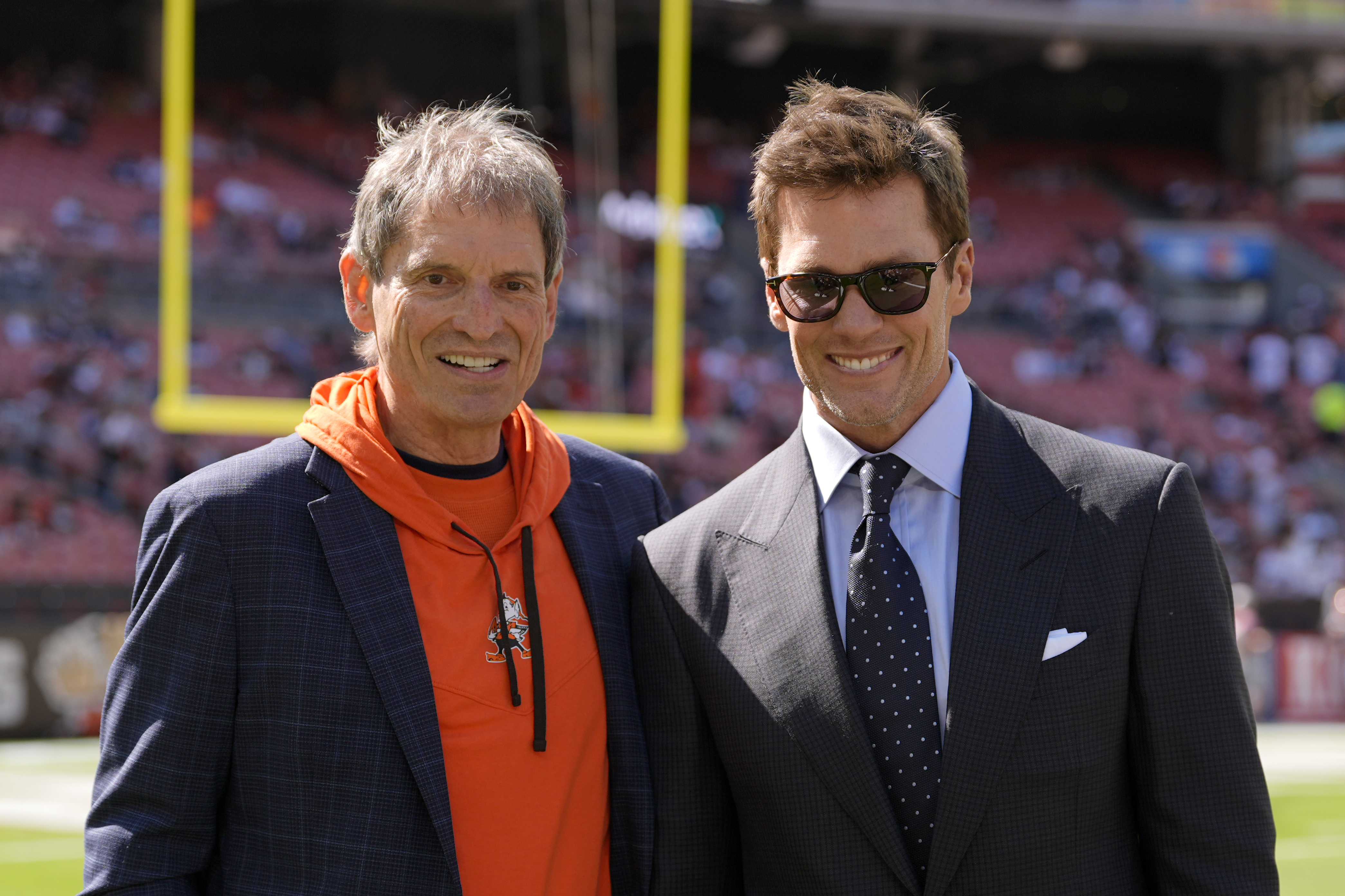 Former NFL players Bernie Kosar, left, and Tom Brady, right, pose for a photo as they talk on the field during warmups before a football game between the Dallas Cowboys and the Cleveland Browns in Cleveland, Sunday, Sept. 8, 2024. (AP Photo/Sue Ogrocki)