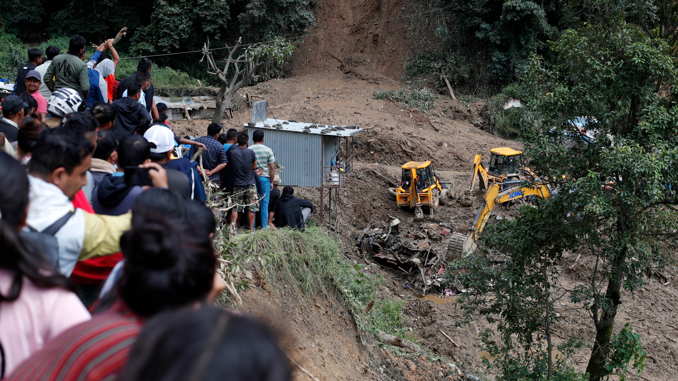 People watch earthmovers removing automobile debris and the dead bodies of victims trapped under a landslide caused by heavy rains in Kathmandu, Nepal, Sunday, Sept. 29, 2024. (AP Photo/Sujan Gurung)