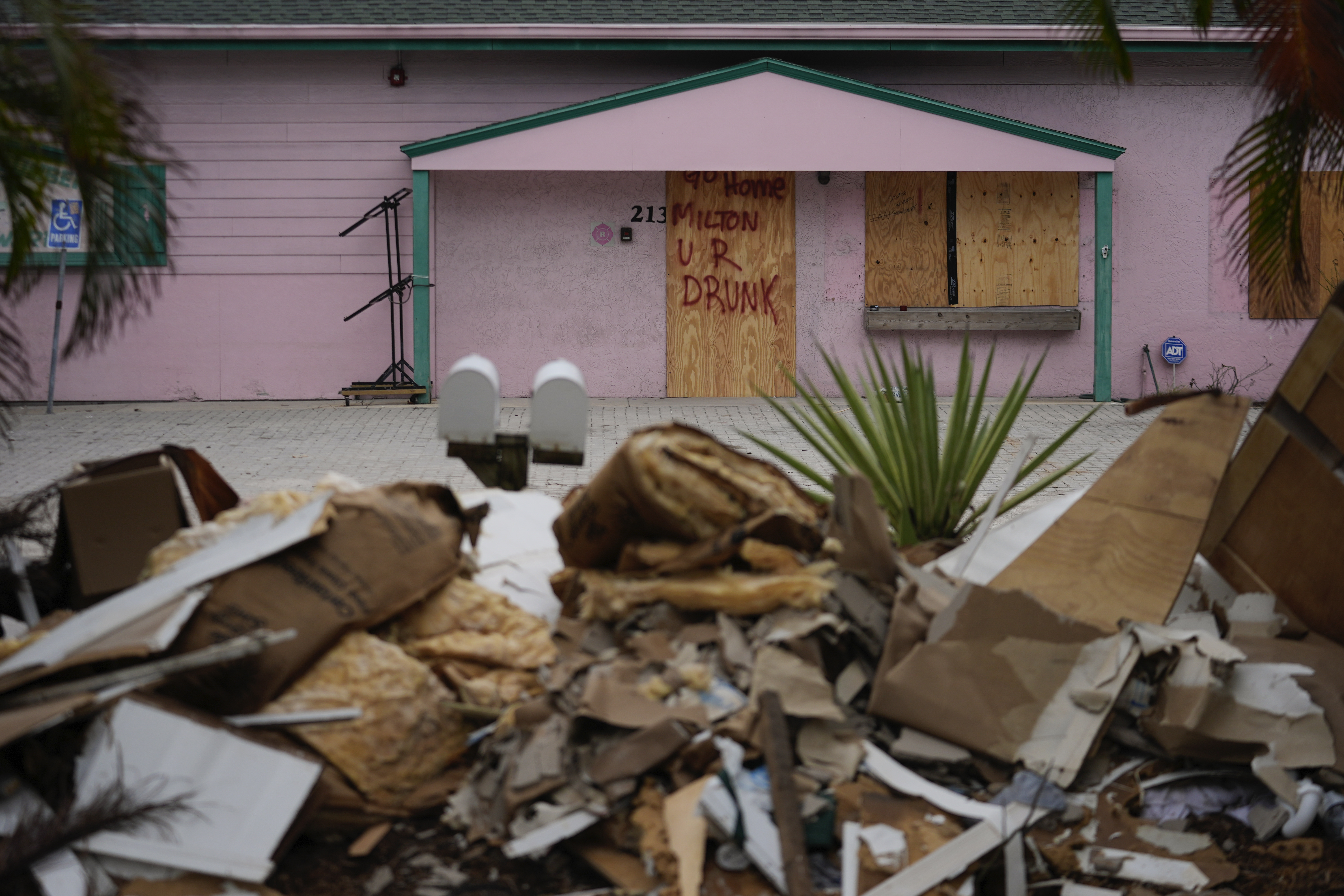 A boarded up business, marked with graffiti reading "Go home Milton, U R drunk," is seen past debris from Hurricane Helene flooding piled up outside a home, ahead of the arrival of Hurricane Milton, in Holmes Beach on Anna Maria Island, Fla., Tuesday, Oct. 8, 2024. (AP Photo/Rebecca Blackwell)