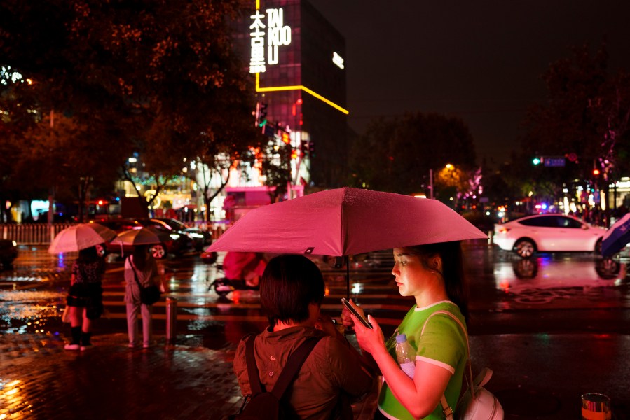 FILE - People look at their phones while waiting to cross an intersection in the rain at the Taikoo Li Sanlitun shopping center in Beijing, July, 30, 2024. (AP Photo/Vincent Thian, File)