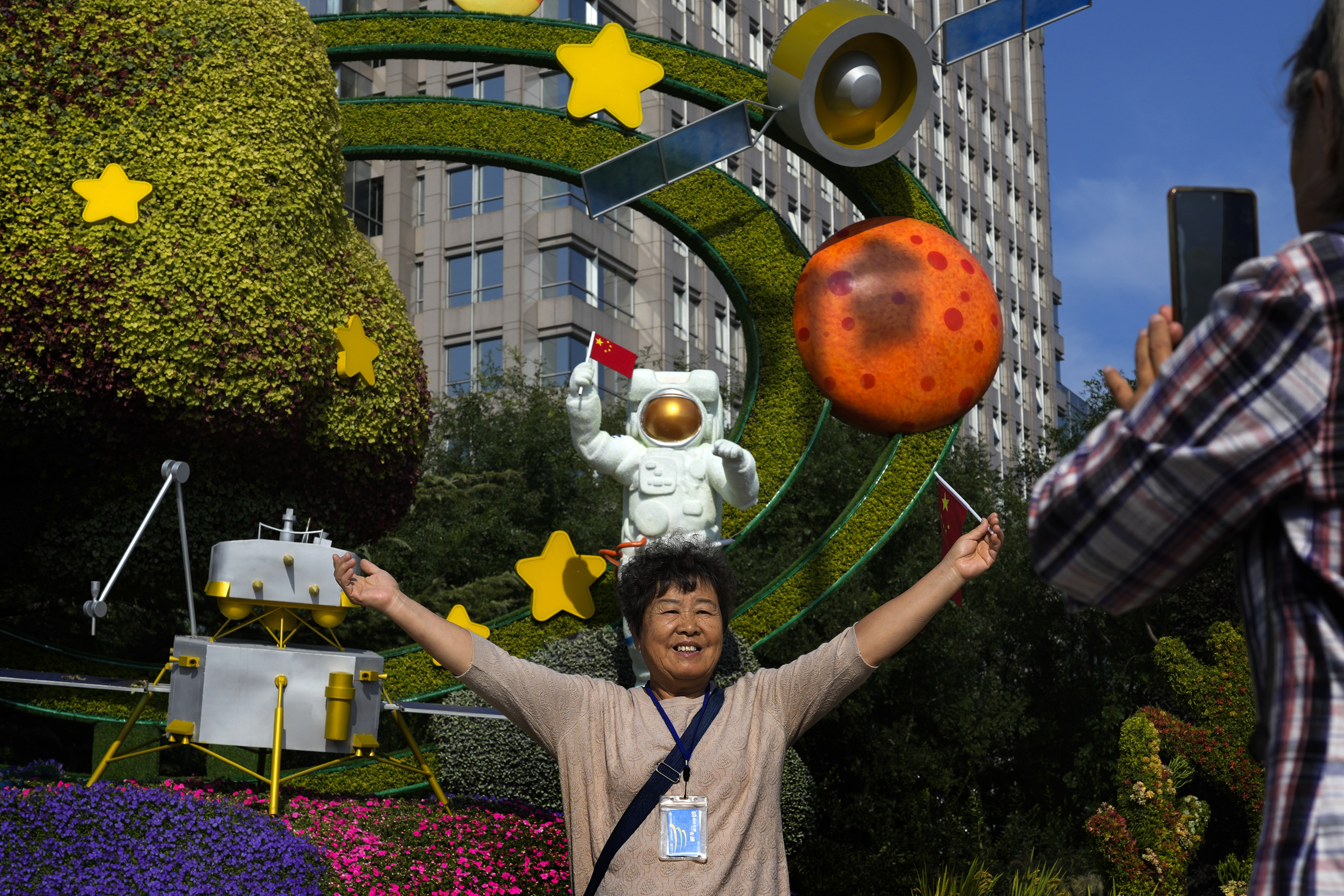 A woman holds a national flag near a floral display featuring a theme of China Space Mission for the upcoming National Day celebration, in Beijing, on Sept. 23, 2024. (AP Photo/Andy Wong, File)