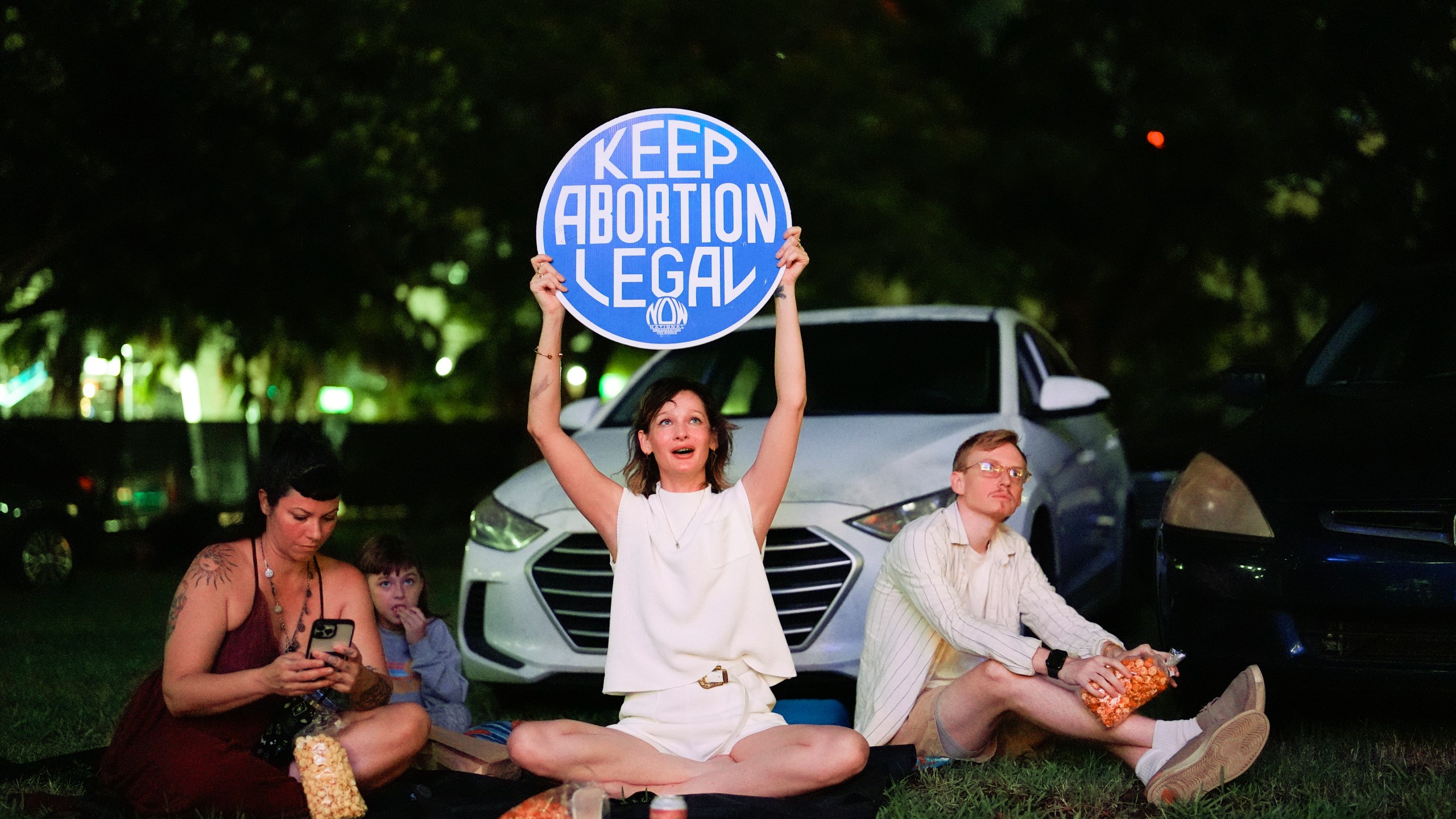 FILE - Reproductive rights advocate Kat Duesterhaus holds up a sign as U.S. President Joe Biden and his Republican rival, former President Donald Trump speak about abortion access, as the the first general election debate of the 2024 season is projected on a outdoor screen at the Nite Owl drive-in theater, Thursday, June 27, 2024, in Miami. (AP Photo/Rebecca Blackwell, File)