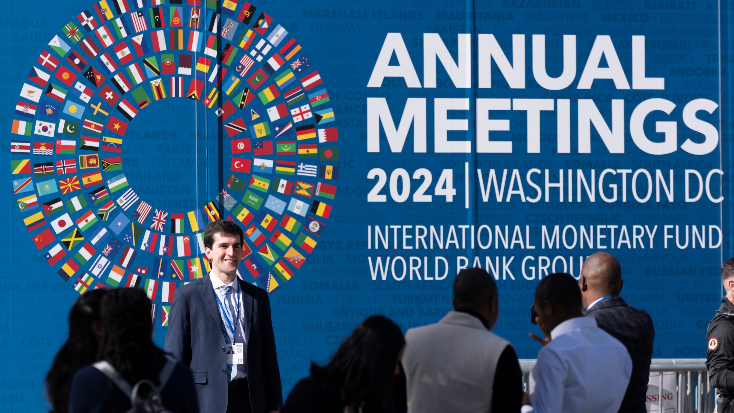 An attendee poses for a photograph in front of a sign advertising the annual meetings of the International Monetary Fund (IMF) and the World Bank Group (WBG), which started in Washington on Monday, Oct. 21, 2024. (AP Photo/Ben Curtis)