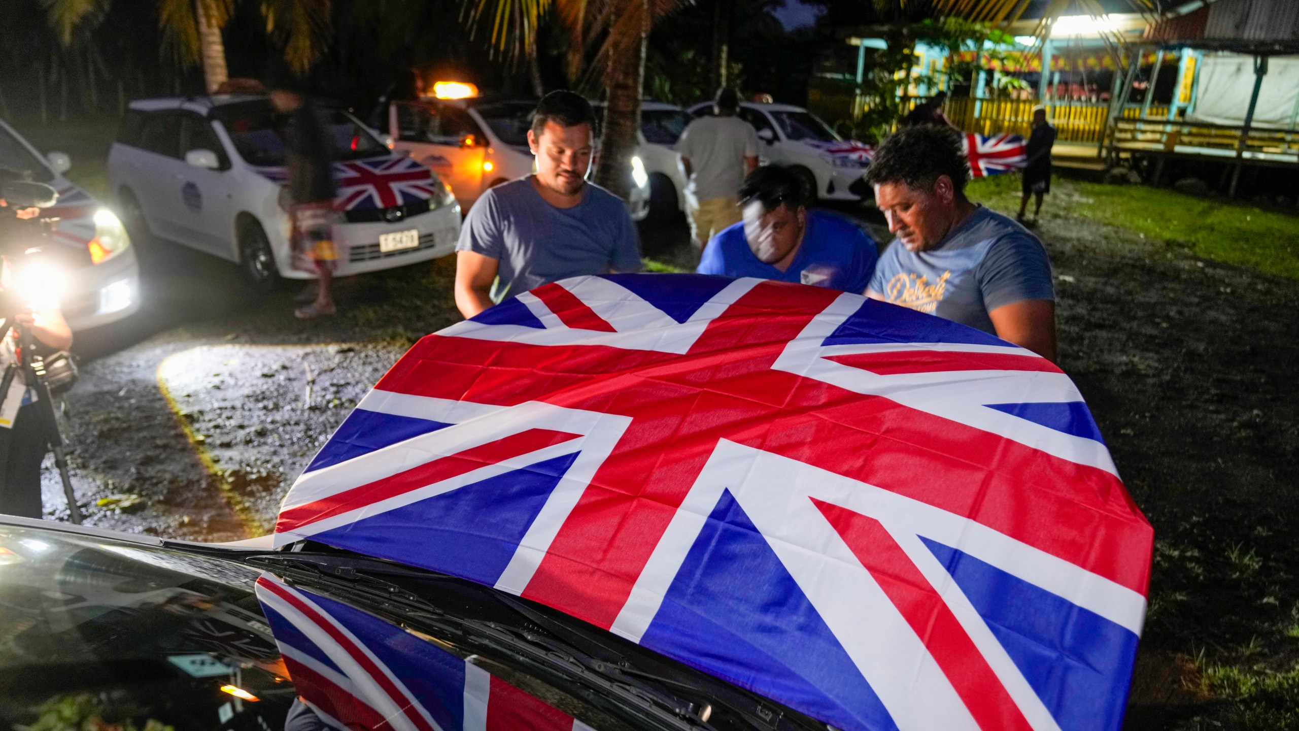 Men attach Union Jacks to the hoods of their taxis as they wait for the arrival of Britain's King Charles III and Queen Camilla in the village of Siumu, Samoa, on Wednesday, Oct. 23, 2024. (AP Photo/Rick Rycroft)