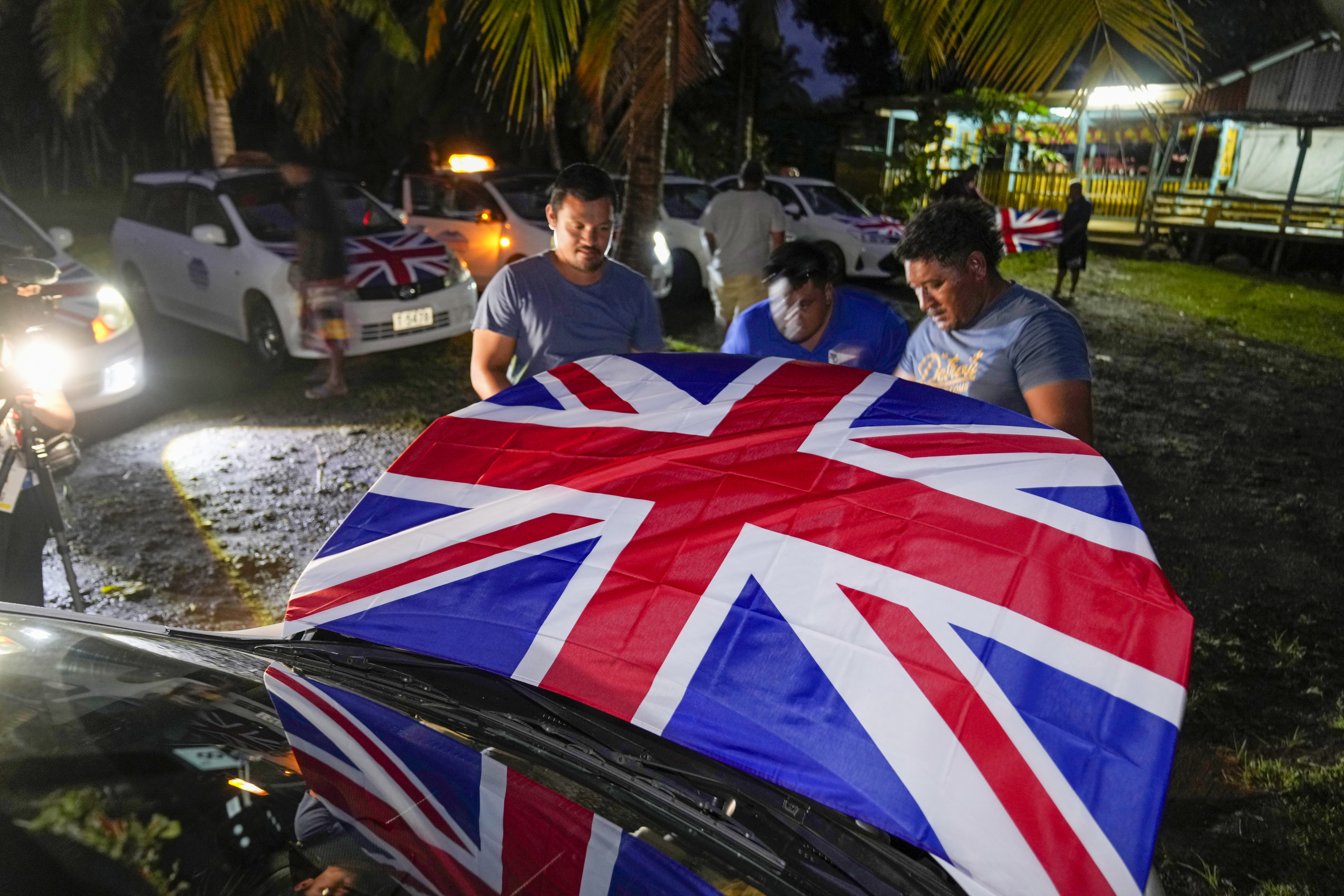 Men attach Union Jacks to the hoods of their taxis as they wait for the arrival of Britain's King Charles III and Queen Camilla in the village of Siumu, Samoa, on Wednesday, Oct. 23, 2024. (AP Photo/Rick Rycroft)