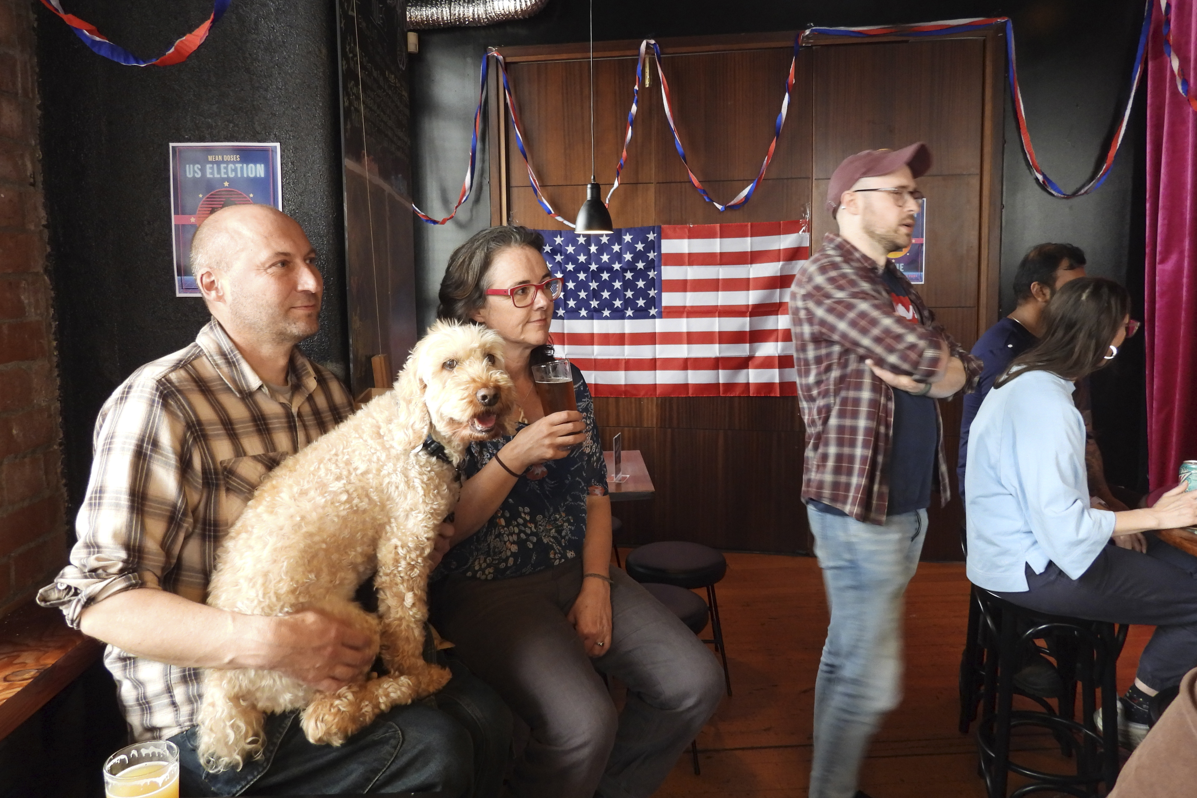 Steve Baker, left, Claudine Earley and their dog Louis watch a television broadcast during a U.S. election viewing party at Mean Doses bar in Wellington, New Zealand on Wednesday, Nov. 6, 2024. (AP Photo/Charlotte Graham-McLay)