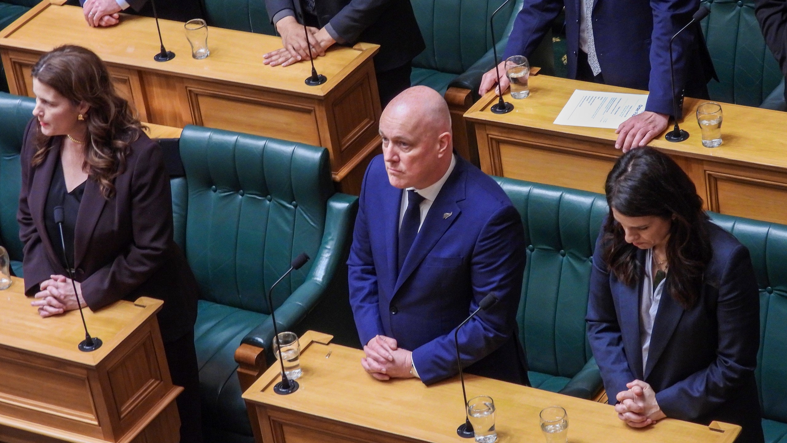 New Zealand Prime Minister Christopher Luxon, center, stands in silence ahead of making a "formal and unreserved" apology in Parliament for the widespread abuse, torture and neglect of hundreds of thousands of children and vulnerable adults in care, in Wellington, New Zealand Tuesday, Nov. 12, 2024. (AP Photo/Charlotte Graham-McLay )