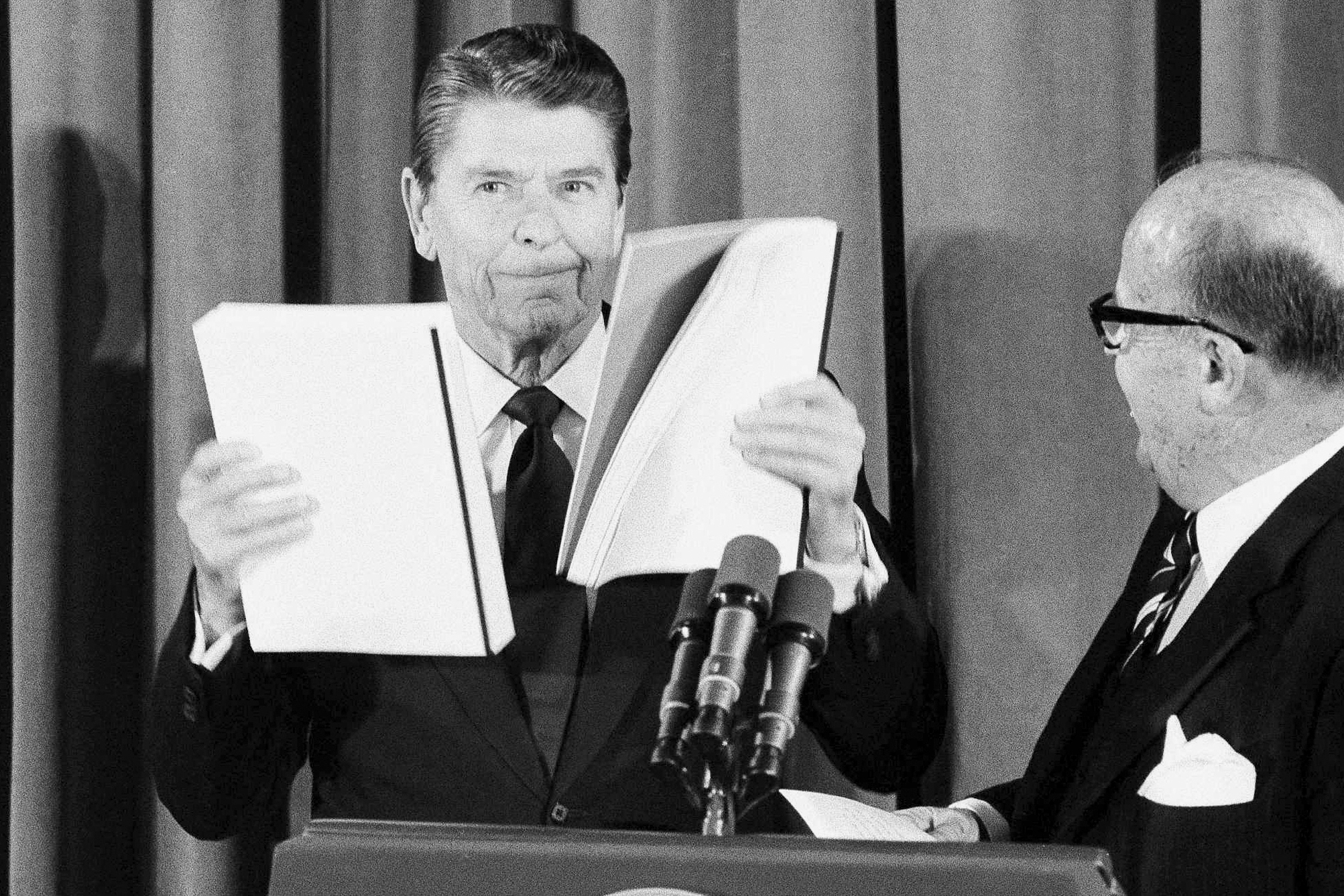 FILE - President Ronald Reagan displays reports given to him by J. Peter Grace, right, of the Private Sector Survey on Waste and Cost Control in Government, at the White House in Washington, Jan 16, 1984. (AP Photo/Ira Schwarz, File)