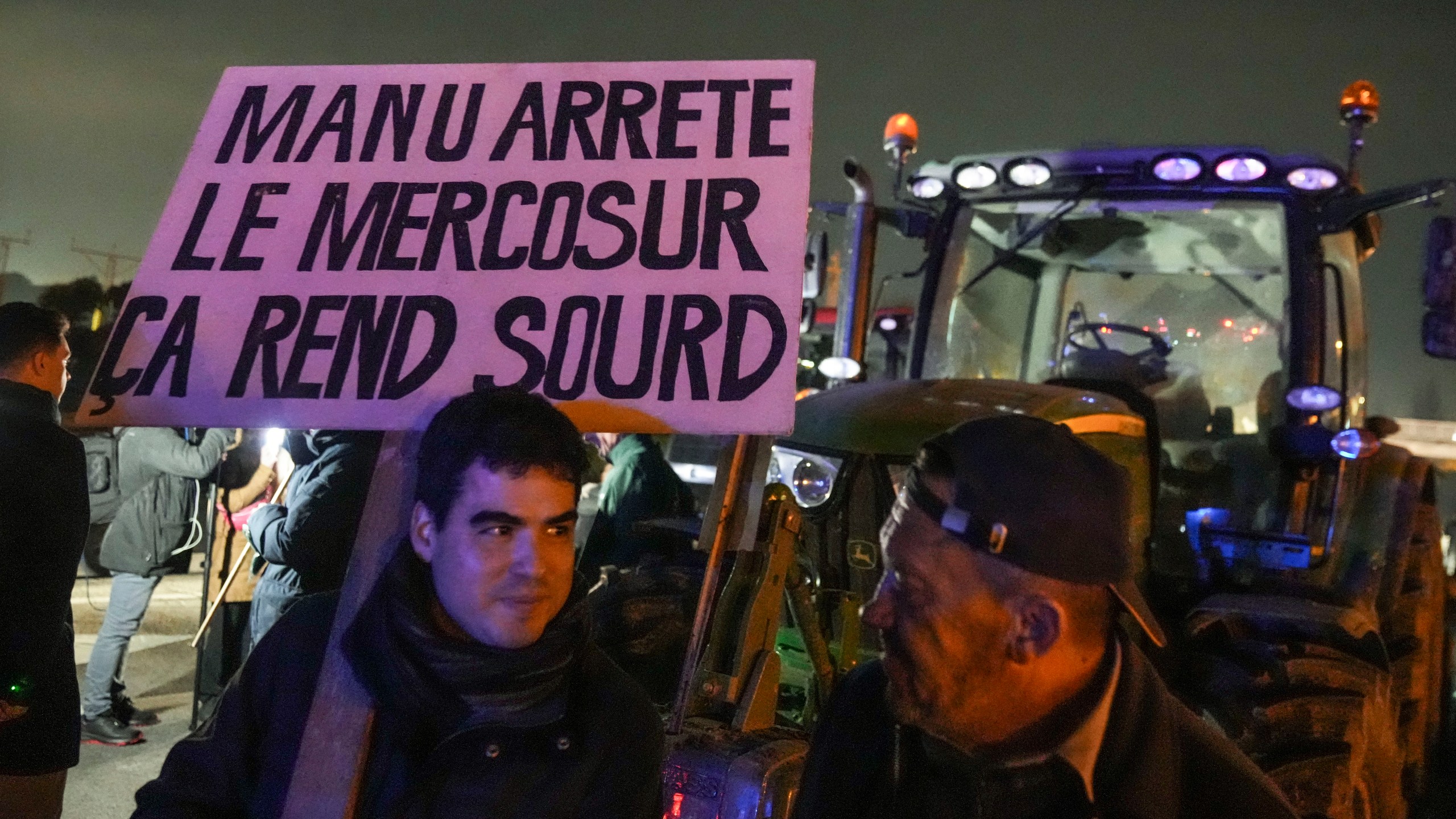 A farmer holds a placard that reads, "Macron stop the Mercosur it makes you deaf " as he stands next to tractors on a blocked highway in Velizy-Villacoublay, outside Paris, Sunday, Nov. 17, 2024. (AP Photo/Michel Euler)