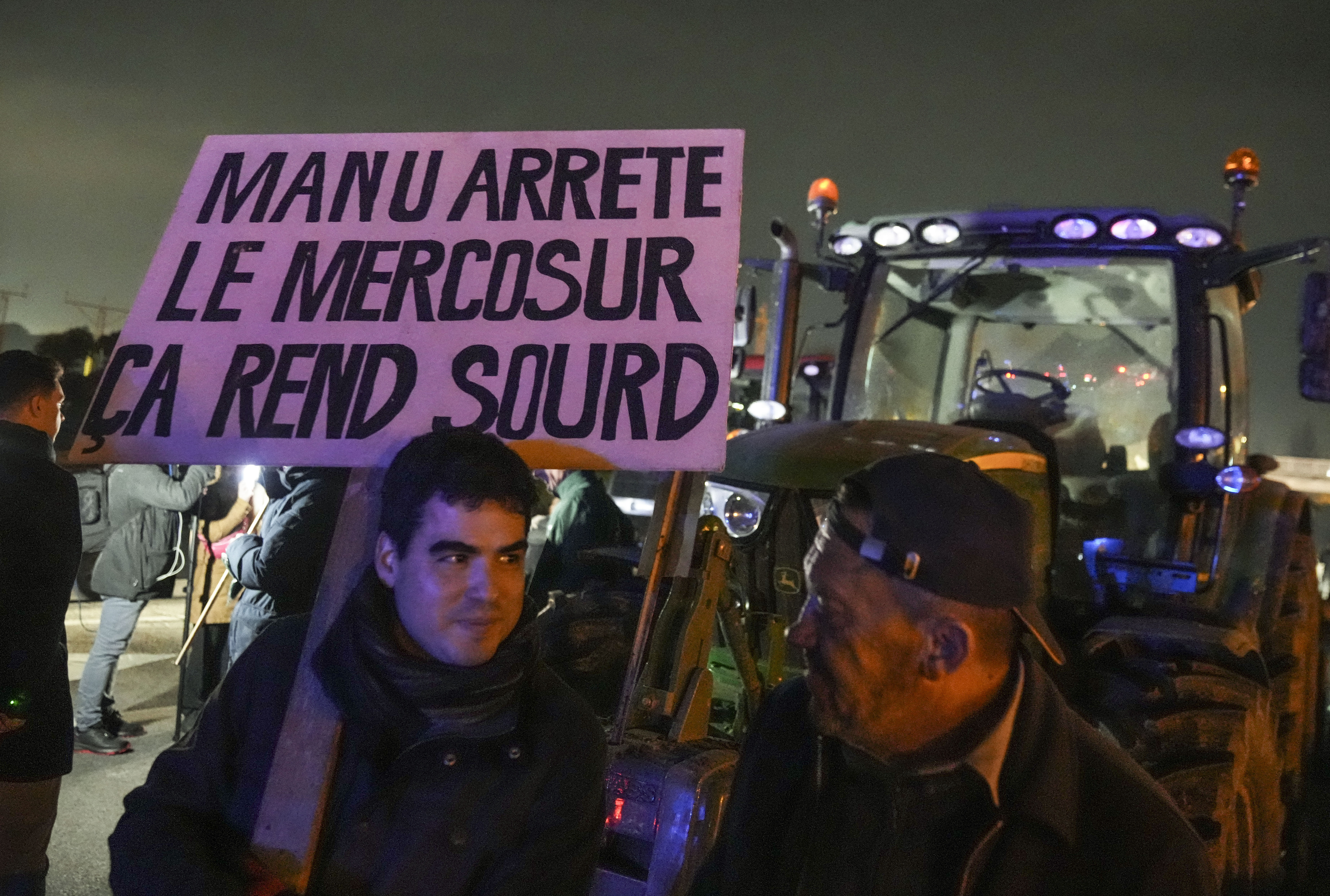 A farmer holds a placard that reads, "Macron stop the Mercosur it makes you deaf " as he stands next to tractors on a blocked highway in Velizy-Villacoublay, outside Paris, Sunday, Nov. 17, 2024. (AP Photo/Michel Euler)