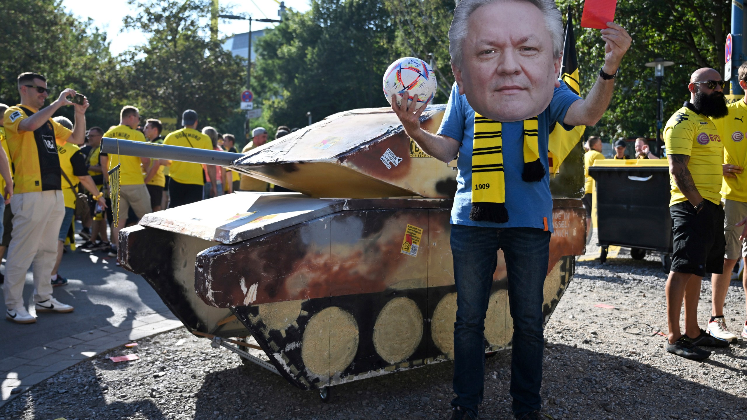 FILE - A dummy tank stands next to an activist wearing a mask with the likeness of Rheinmetall CEO Armin Papperger and holding up a red card ahead of the soccer game between Borussia Dortmund and Eintracht Frankfurt, in Dortmund, Germany, Saturday Aug. 24, 2024. (Bernd Thissen/dpa via AP, File)