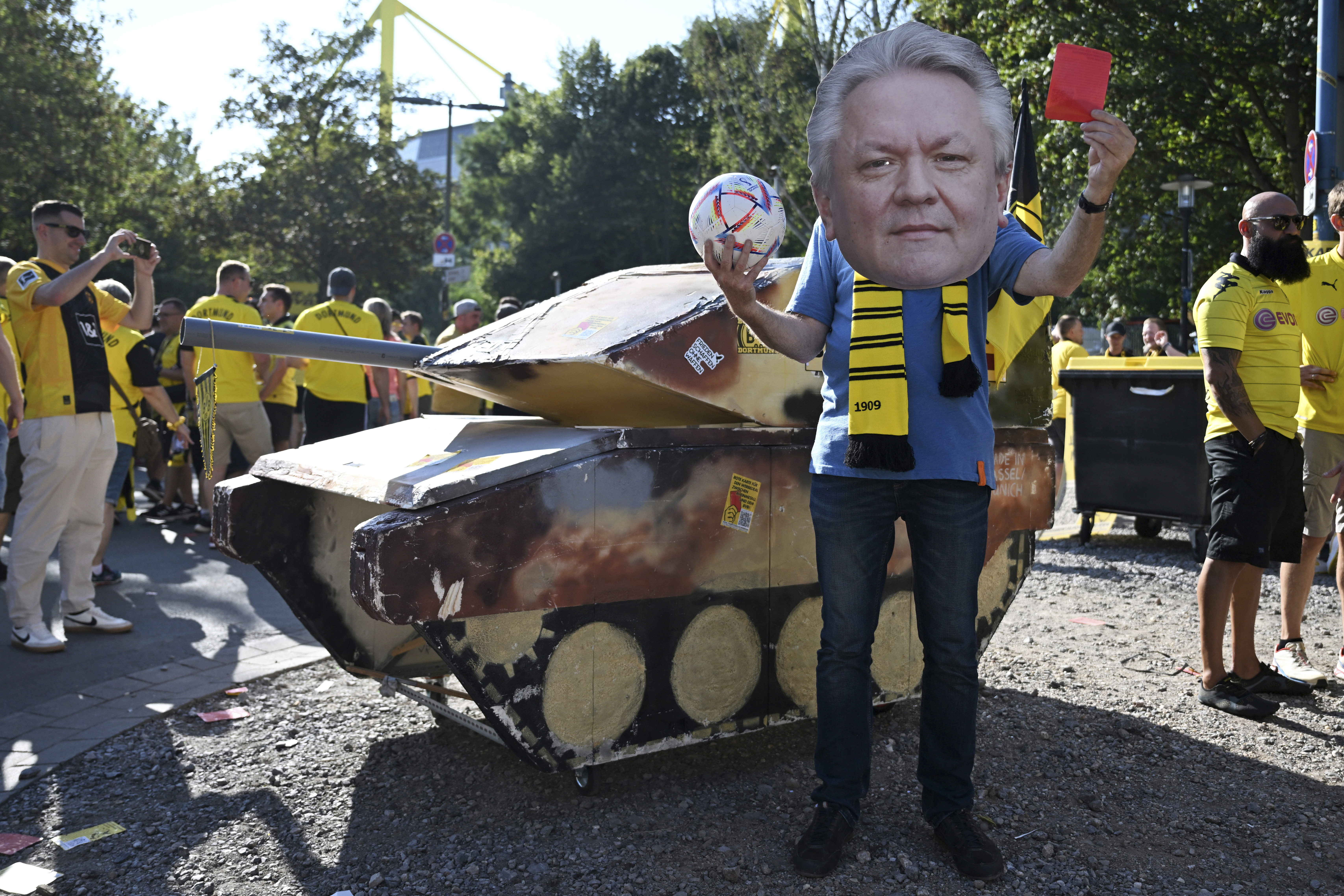 FILE - A dummy tank stands next to an activist wearing a mask with the likeness of Rheinmetall CEO Armin Papperger and holding up a red card ahead of the soccer game between Borussia Dortmund and Eintracht Frankfurt, in Dortmund, Germany, Saturday Aug. 24, 2024. (Bernd Thissen/dpa via AP, File)