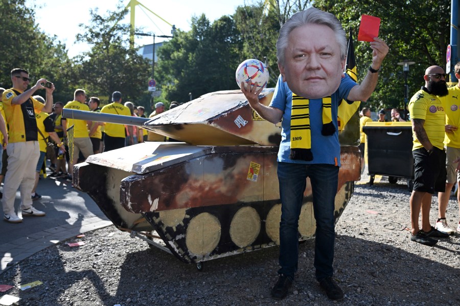 FILE - A dummy tank stands next to an activist wearing a mask with the likeness of Rheinmetall CEO Armin Papperger and holding up a red card ahead of the soccer game between Borussia Dortmund and Eintracht Frankfurt, in Dortmund, Germany, Saturday Aug. 24, 2024. (Bernd Thissen/dpa via AP, File)