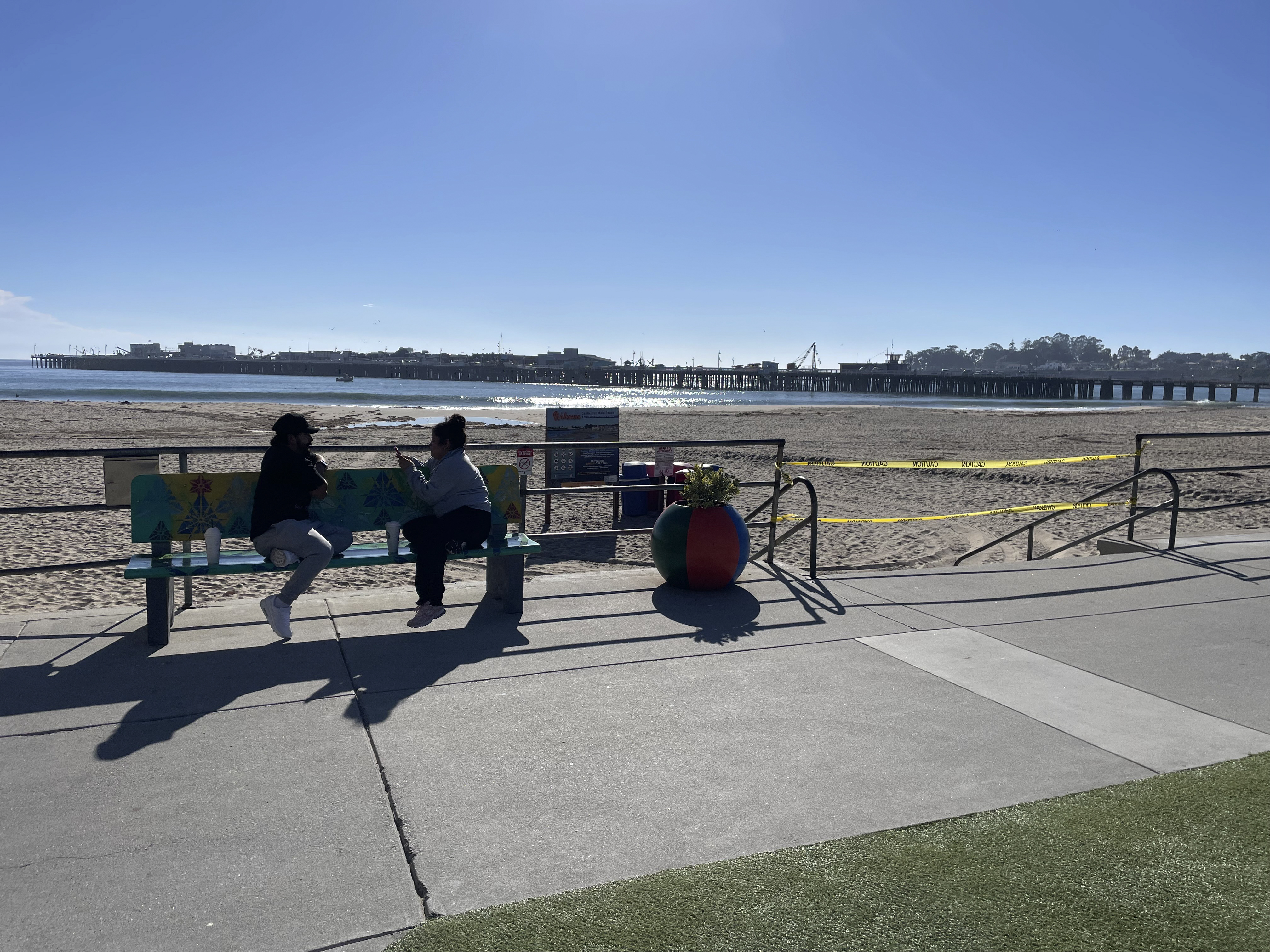 People sit on a bench along the beach in Santa Cruz, Calif. after authorities cleared the main beach, taping off entrances on Thursday, Dec. 5, 2024. (AP Photo/Martha Mendoza)