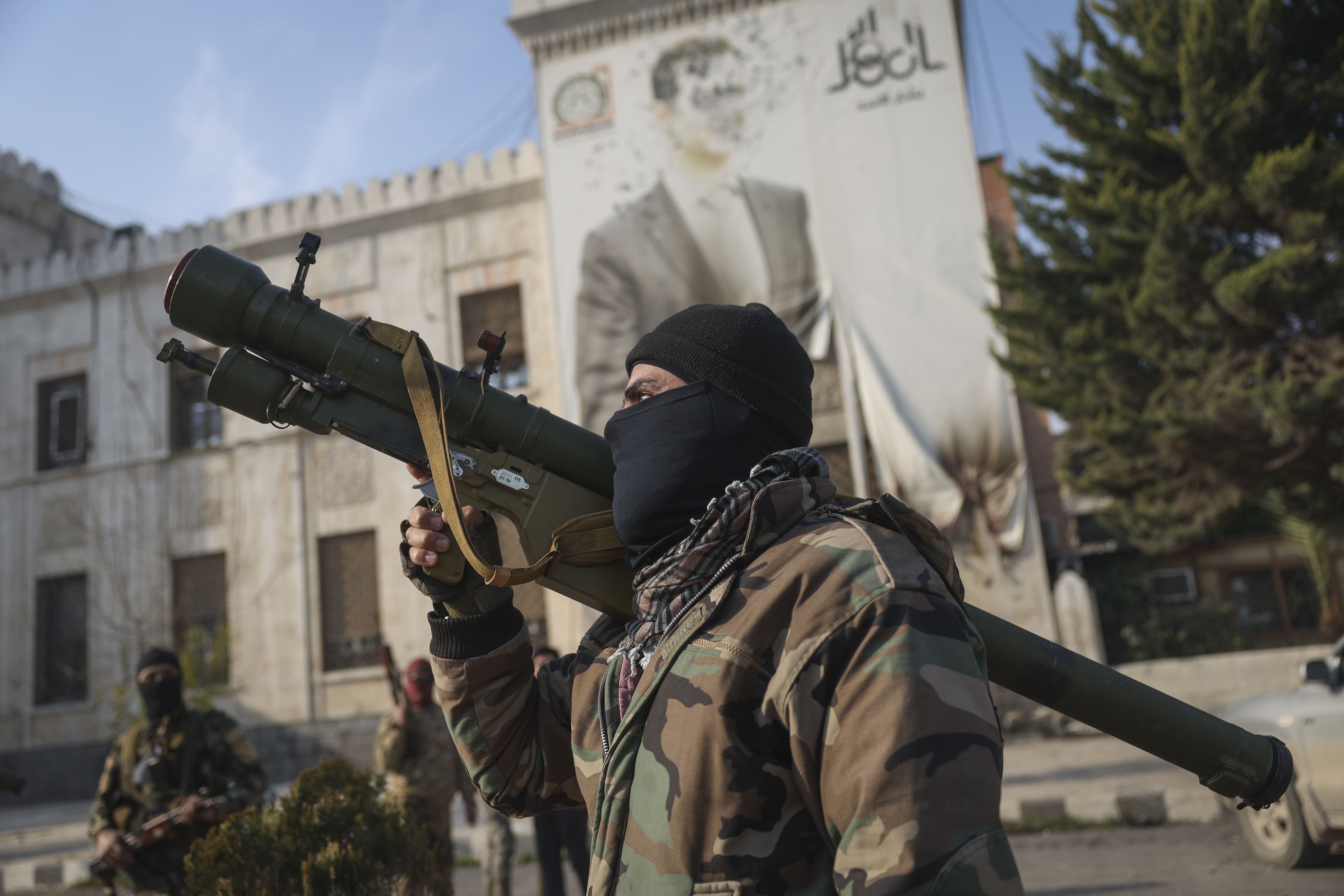 A Syrian opposition fighter holds a rocket launcher in front of the provincial government office, where an image of Syrian President Bashar Assad is riddled with bullets on the facade, in the aftermath of the opposition's takeover of Hama, Syria, Friday, Dec. 6, 2024. (AP Photo/Ghaith Alsayed)