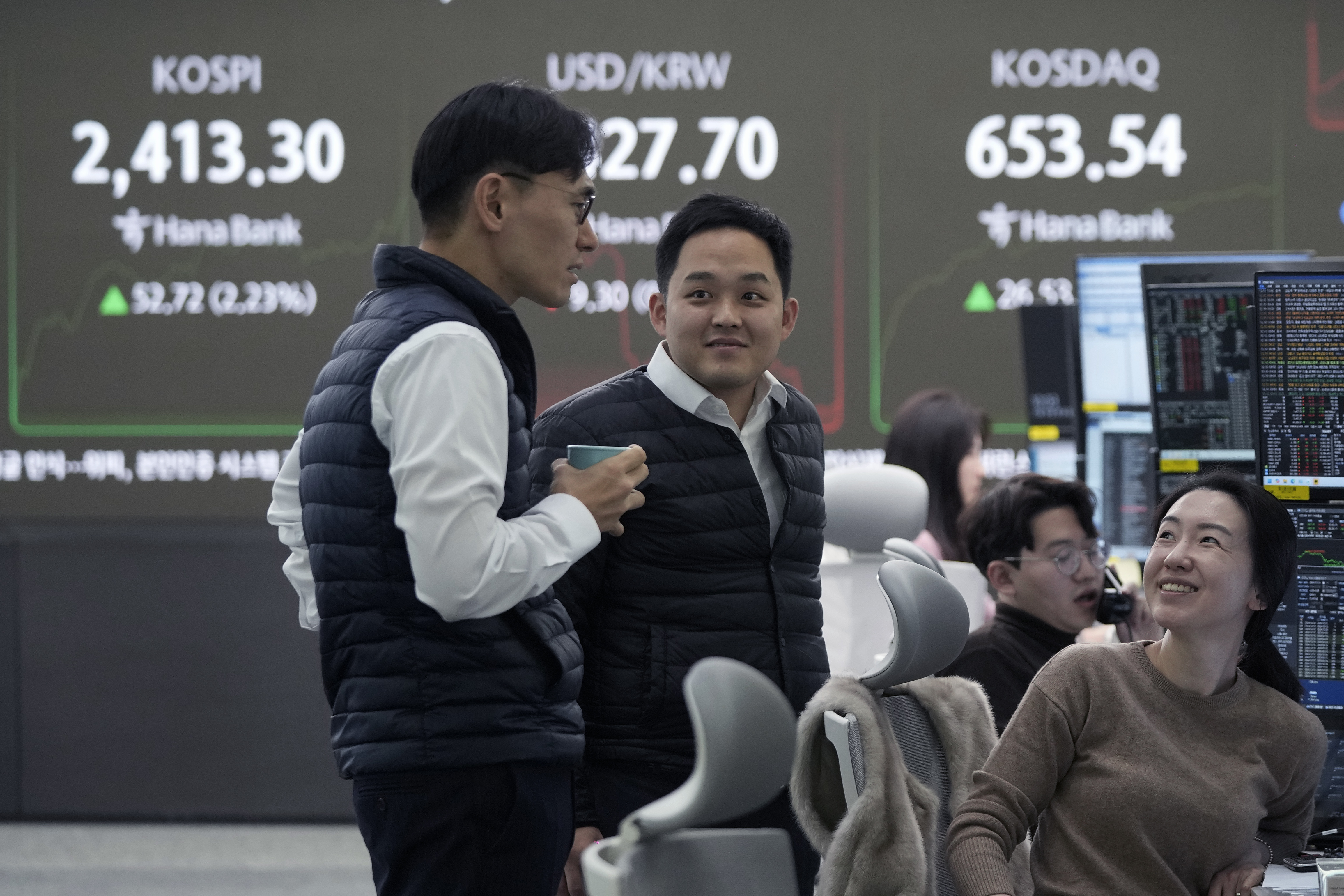 Currency traders smile near a screen showing the Korea Composite Stock Price Index (KOSPI), top left, and the foreign exchange rate between U.S. dollar and South Korean won, top center, at the foreign exchange dealing room of the KEB Hana Bank headquarters in Seoul, South Korea, Tuesday, Dec. 10, 2024. (AP Photo/Ahn Young-joon)