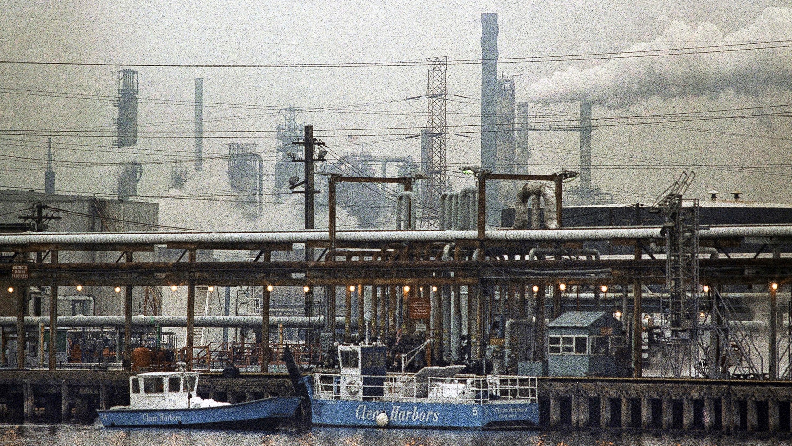 FILE - The Exxon oil refinery stands in Linden, N.J., Jan. 9, 1990, as two clean harbor cleanup boats work in the Arthur Kill waterway. (AP Photo/Mike Derer, File)