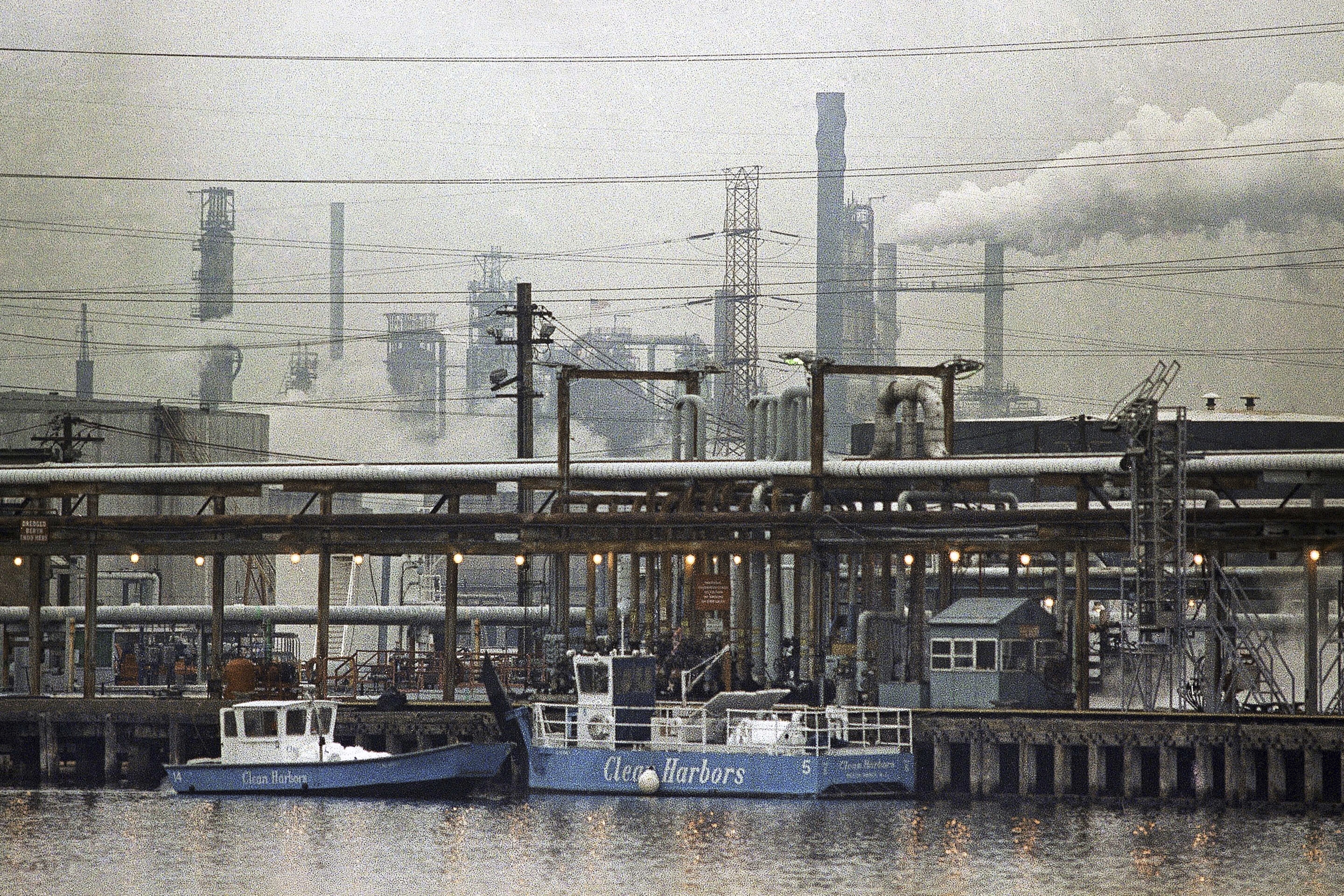 FILE - The Exxon oil refinery stands in Linden, N.J., Jan. 9, 1990, as two clean harbor cleanup boats work in the Arthur Kill waterway. (AP Photo/Mike Derer, File)