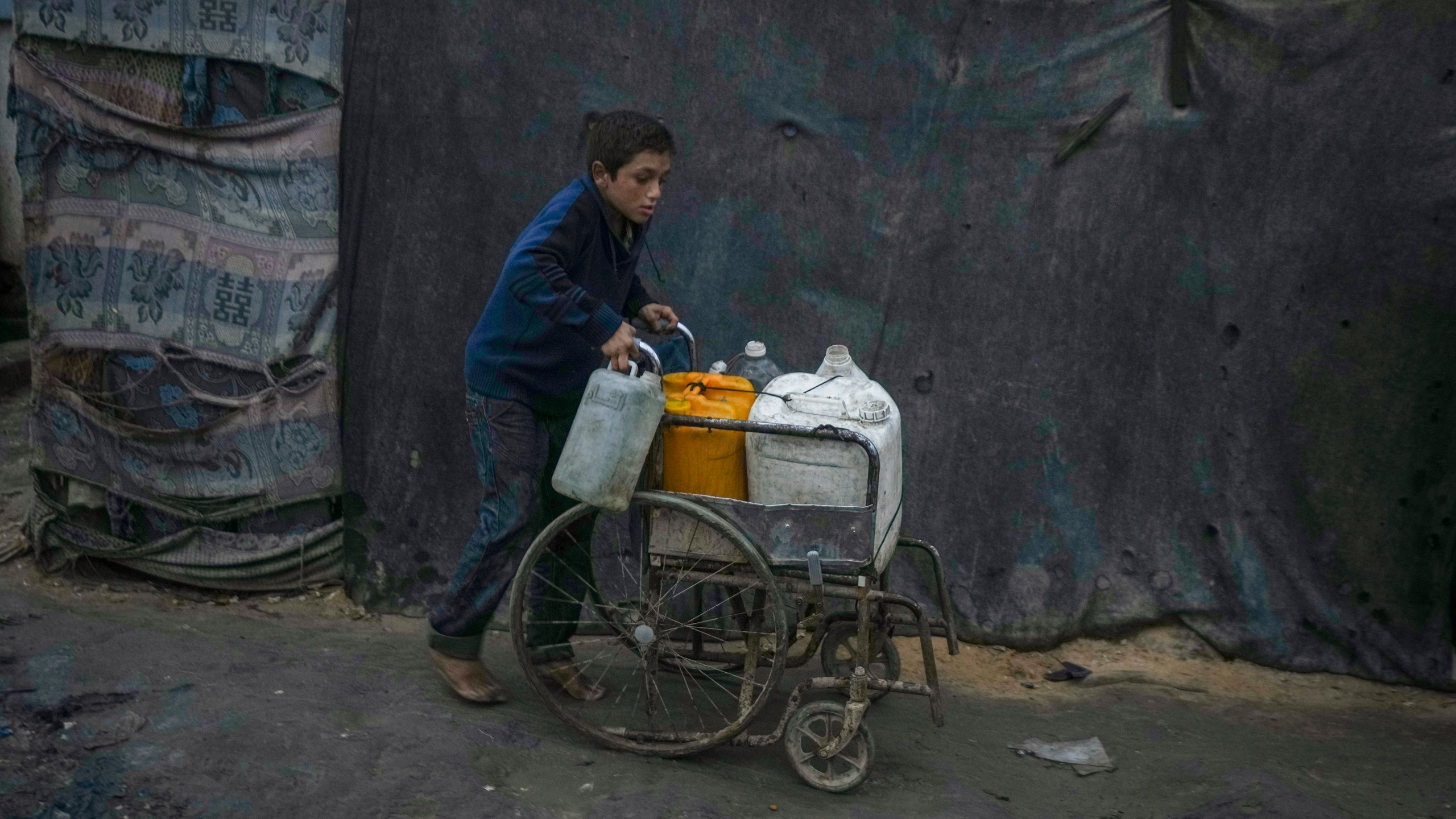 A Palestinian boy pushes a wheelchair carrying jerrycans and plastic bottles with water at a camp for displaced people in Deir al-Balah, Gaza Strip, Thursday, Dec. 12, 2024. (AP Photo/Abdel Kareem Hana)