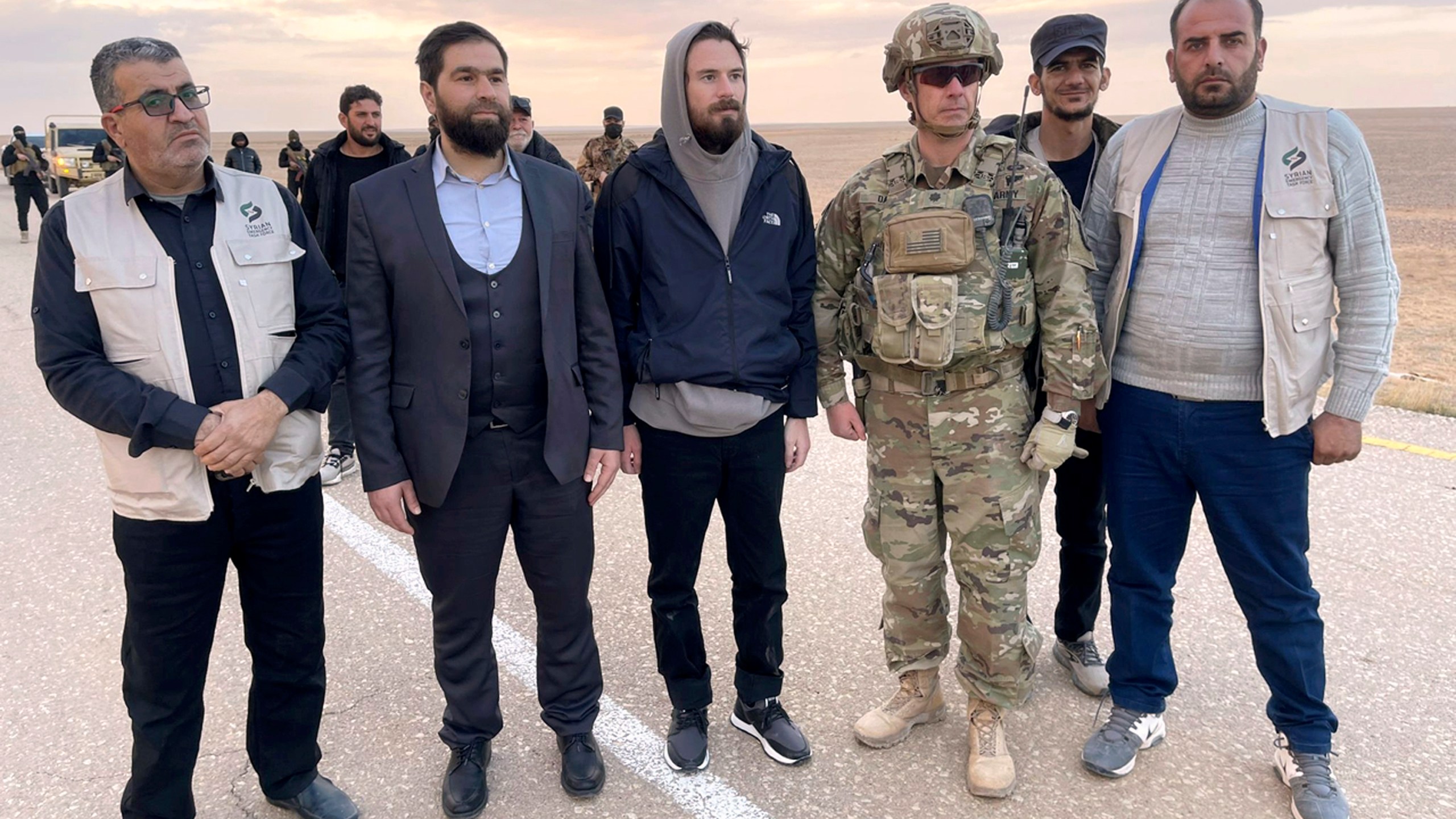 This image provided by Mouaz Moustafa shows Travis Timmerman, center, standing with a member of the U.S. military and representatives of Syria's transitional government and opposition activists near the Syria-Jordanian border Friday, Dec. 13, 2024. U.S. forces flew Timmerman out of Syria Friday after rebels freed him following seven months in a Syrian government prison. (Mouaz Moustafa via AP)