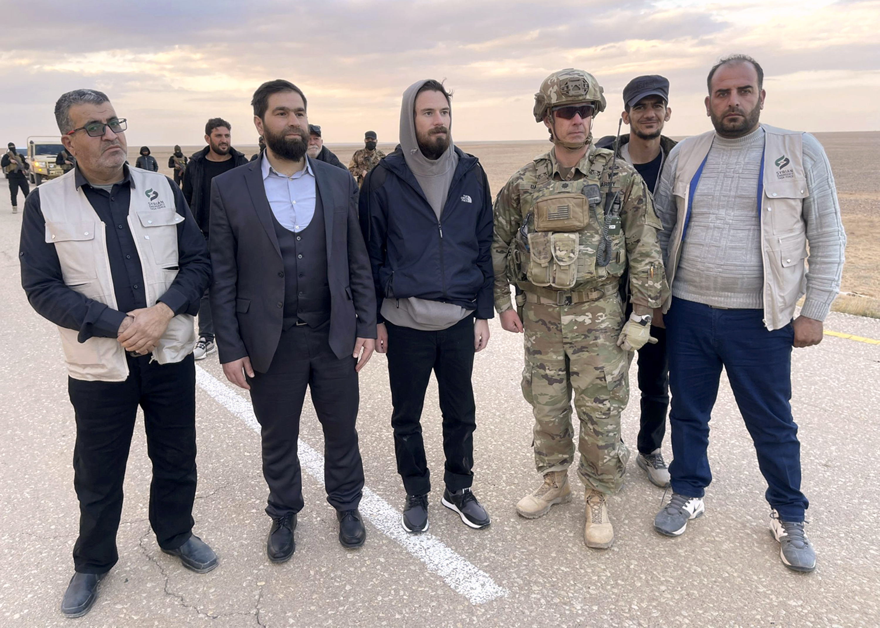 This image provided by Mouaz Moustafa shows Travis Timmerman, center, standing with a member of the U.S. military and representatives of Syria's transitional government and opposition activists near the Syria-Jordanian border Friday, Dec. 13, 2024. U.S. forces flew Timmerman out of Syria Friday after rebels freed him following seven months in a Syrian government prison. (Mouaz Moustafa via AP)