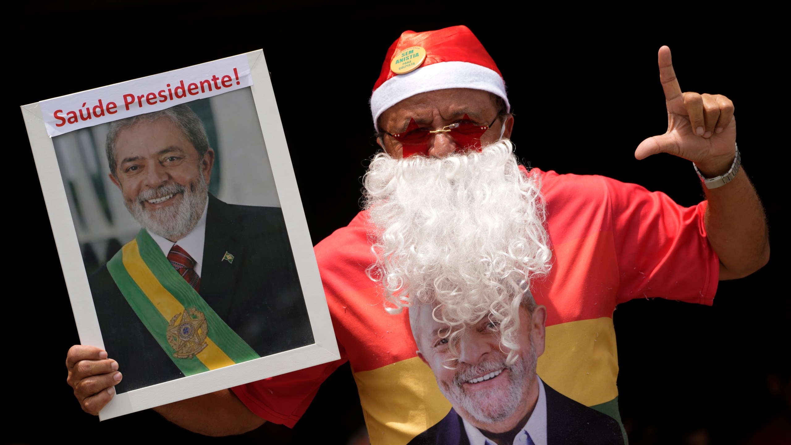 A supporter of Brazilian President Luiz Inacio Lula da Silva dressed as Santa Claus signals the letter "L" for Lula, with a photo that reads in Portuguese "Health President!," during a gathering of support for the leader in Brasilia, Brazil, Tuesday, Dec. 10, 2024, while Lula is hospitalized in Sao Paulo. (AP Photo/Eraldo Peres)