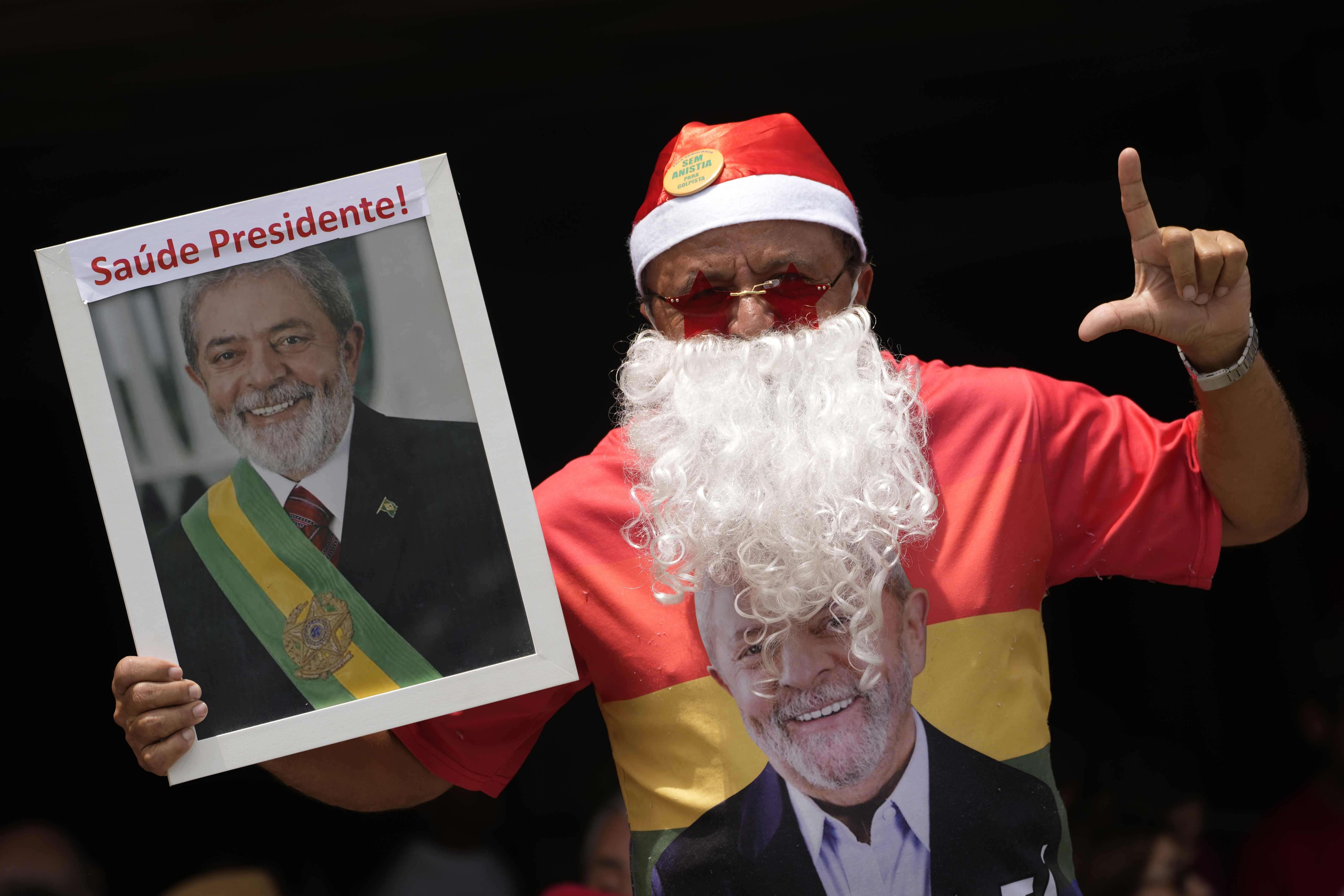 A supporter of Brazilian President Luiz Inacio Lula da Silva dressed as Santa Claus signals the letter "L" for Lula, with a photo that reads in Portuguese "Health President!," during a gathering of support for the leader in Brasilia, Brazil, Tuesday, Dec. 10, 2024, while Lula is hospitalized in Sao Paulo. (AP Photo/Eraldo Peres)