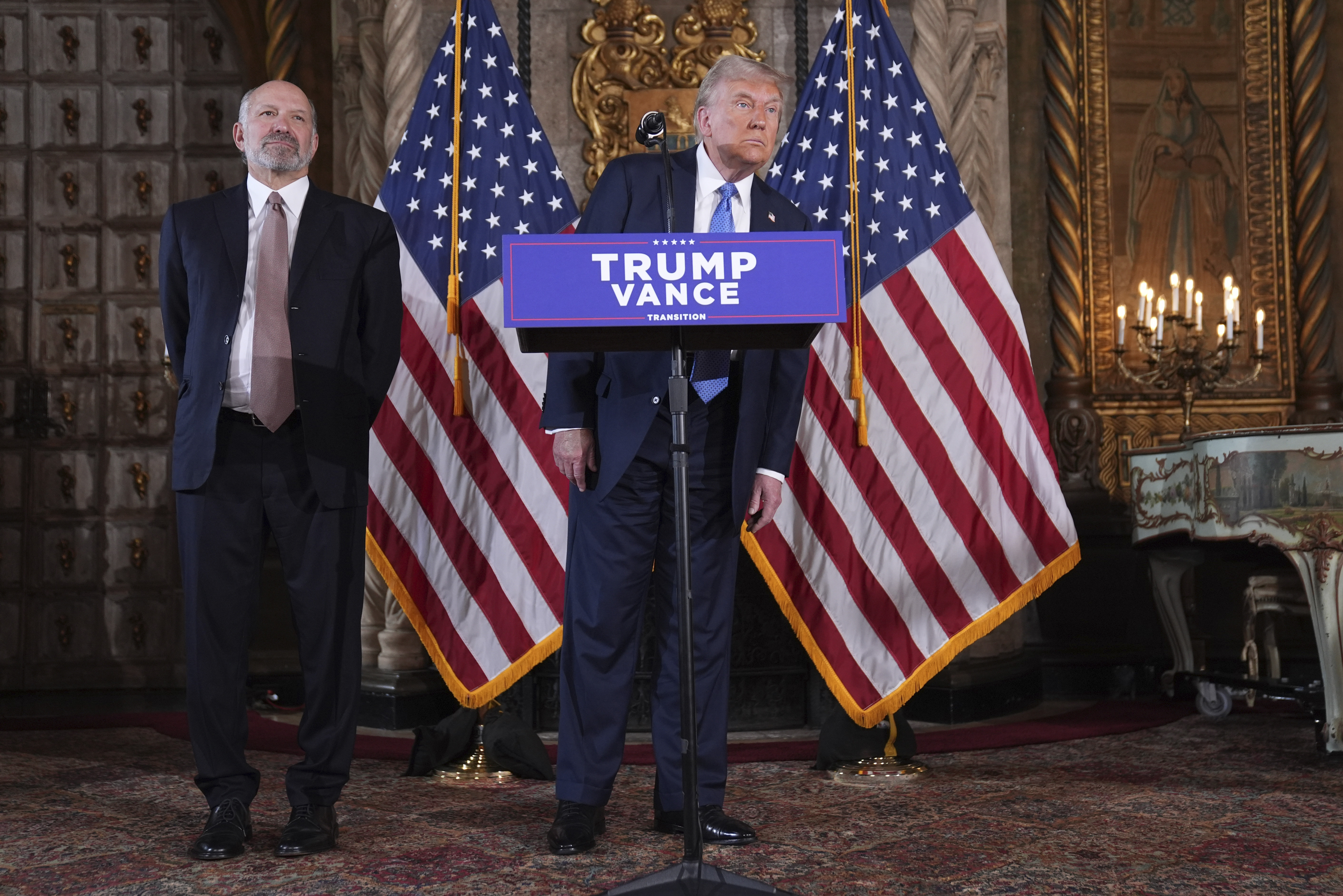 President-elect Donald Trump speaks at a news conference at Mar-a-Lago, Monday, Dec. 16, 2024, in Palm Beach, Fla., as Commerce Secretary nominee Howard Lutnick listens. (AP Photo/Evan Vucci)