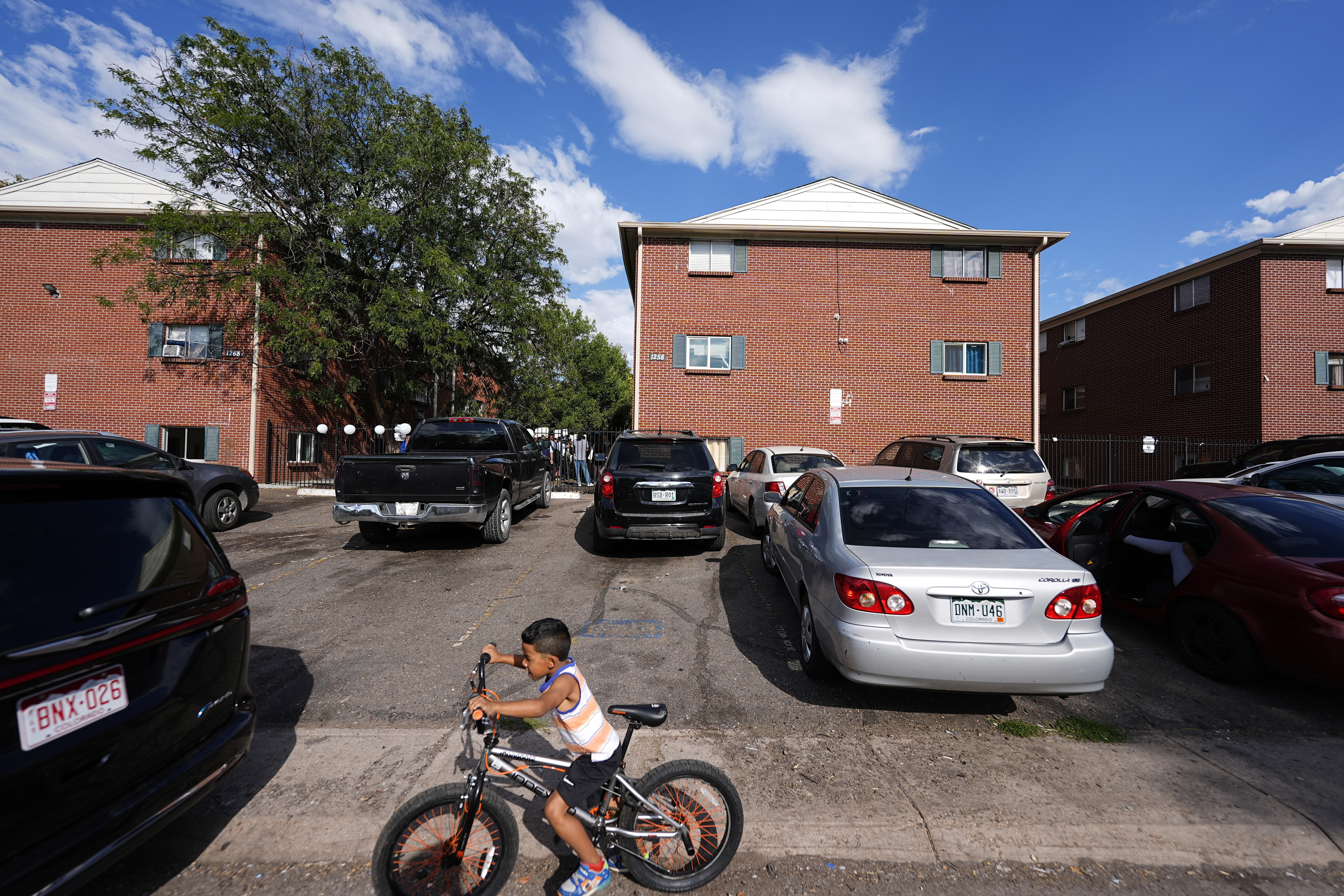 FILE - A boy rides his bicycle past apartment buildings as a rally staged by the East Colfax Community Collective is held in the courtyard to address chronic problems in the apartment buildings occupied by people displaced from their home countries in central and South America, Sept. 3, 2024, in Aurora, Colo. (AP Photo/David Zalubowski, File)