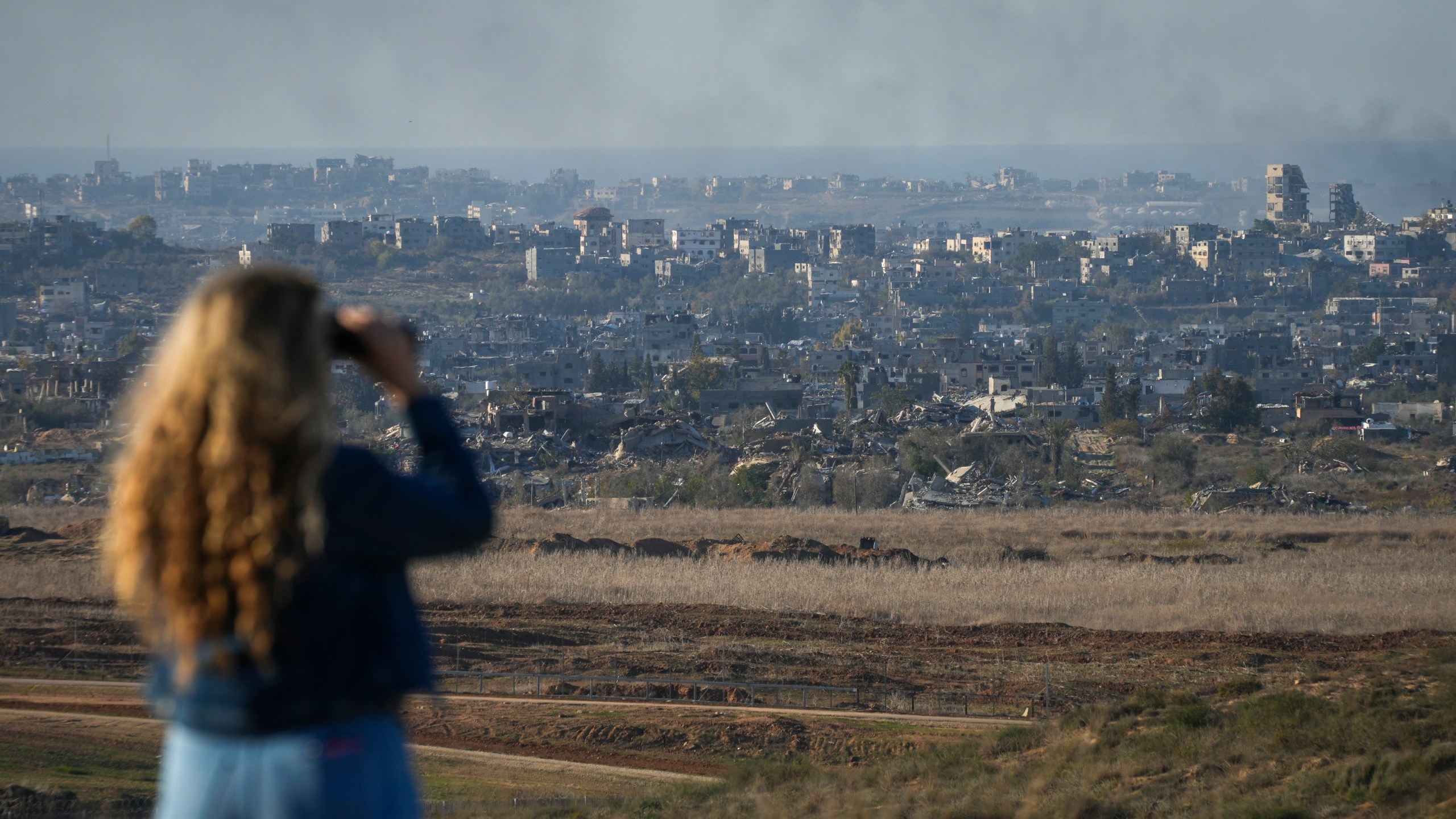 A woman uses binoculars to watch smoke rising following an explosion in the Gaza Strip as seen from southern Israel, Wednesday, Dec.18, 2024. (AP Photo/Ohad Zwigenberg)