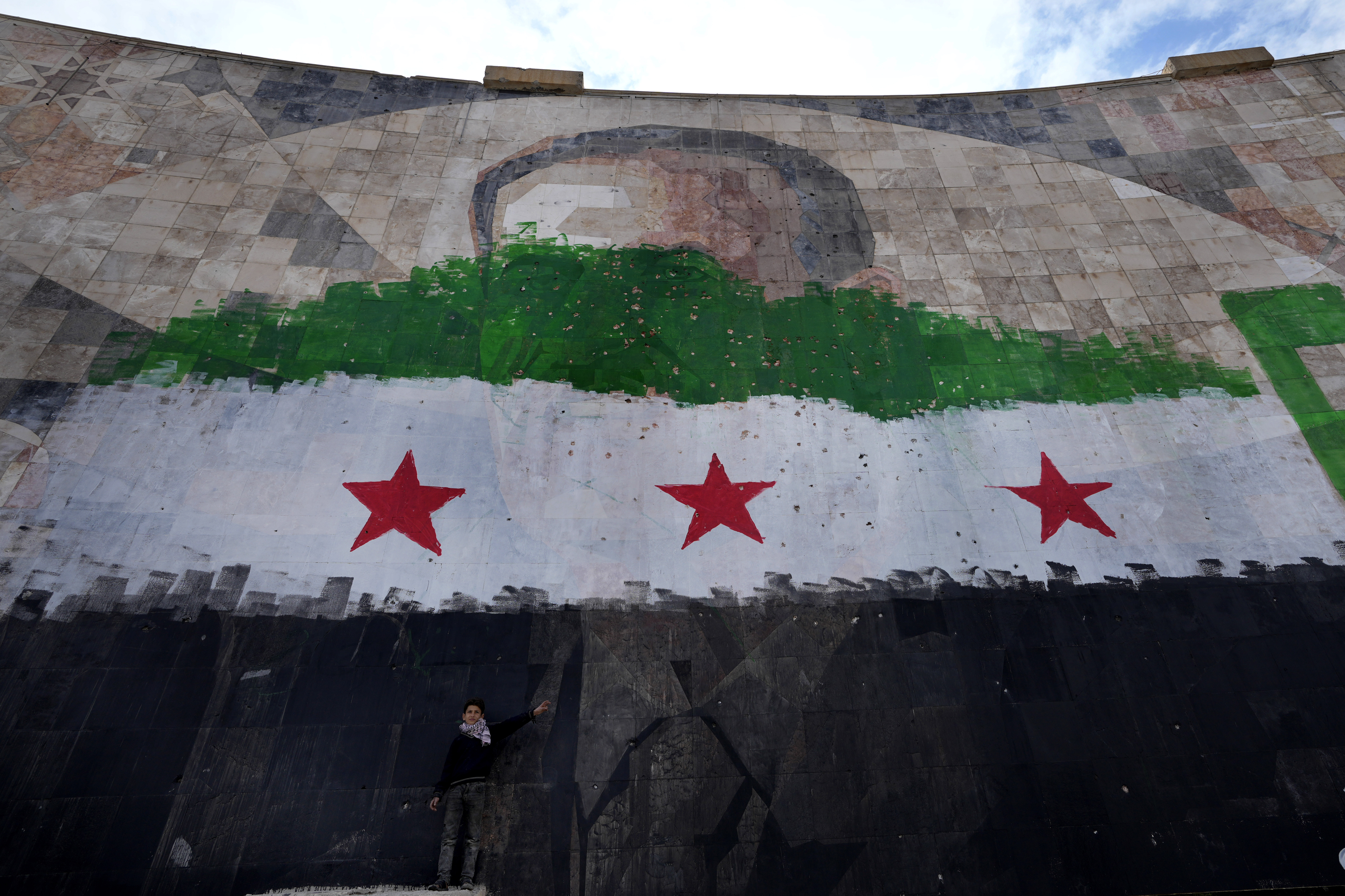 A Syrian boy stands under a giant portrait of the late Syrian President Hafez Assad painted over with the colors of the "revolutionary" flag, in Damascus, Syria, Thursday, Dec. 26, 2024. (AP Photo/Hussein Malla)