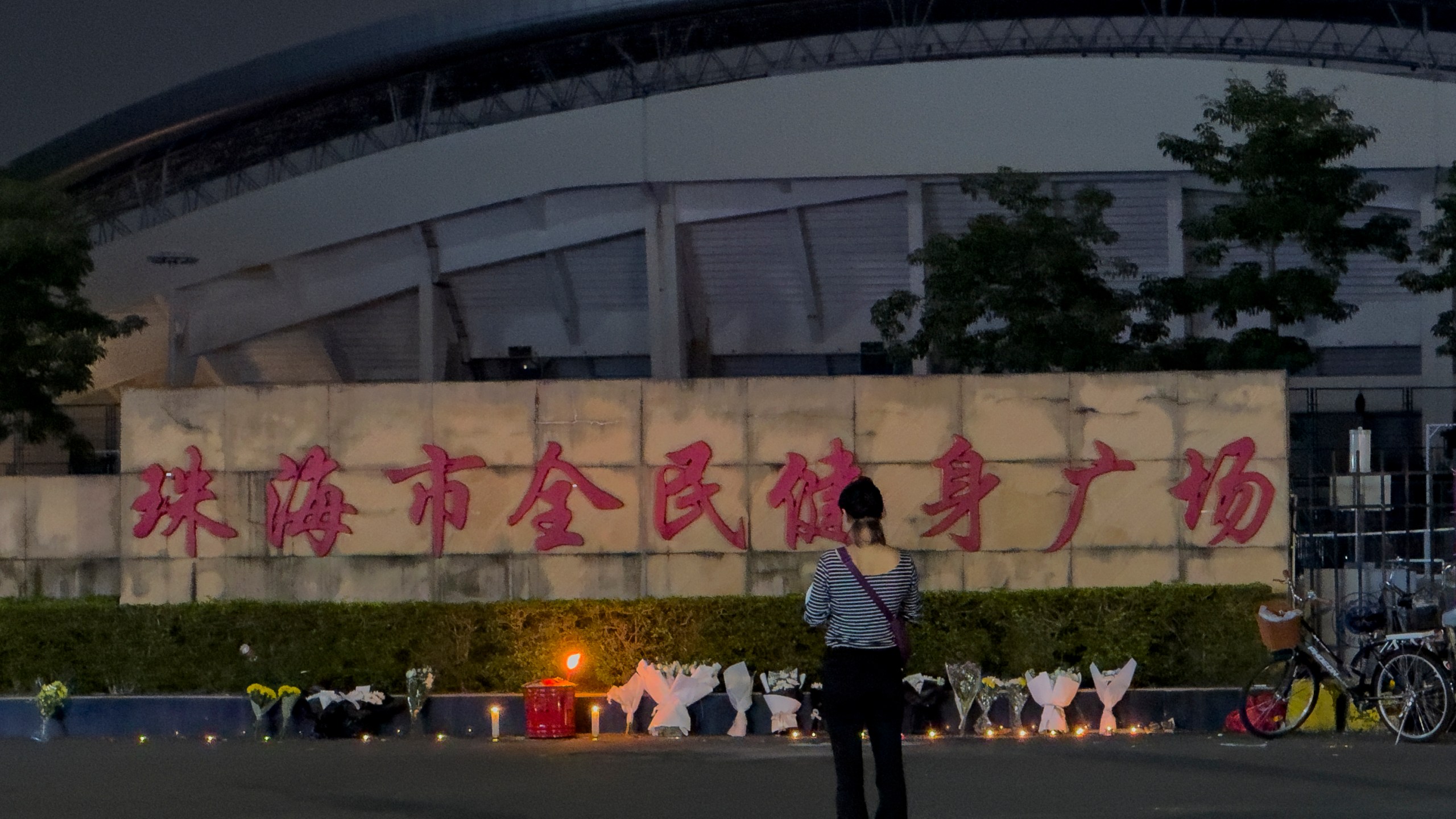 FILE - A woman stands near flowers placed outside the "Zhuhai People's Fitness Plaza", where a man deliberately rammed his car into people exercising at the sports center, killing some and injuring others in Zhuhai in southern China's Guangdong province, Nov. 12, 2024. (AP Photo/Ng Han Guan, File)