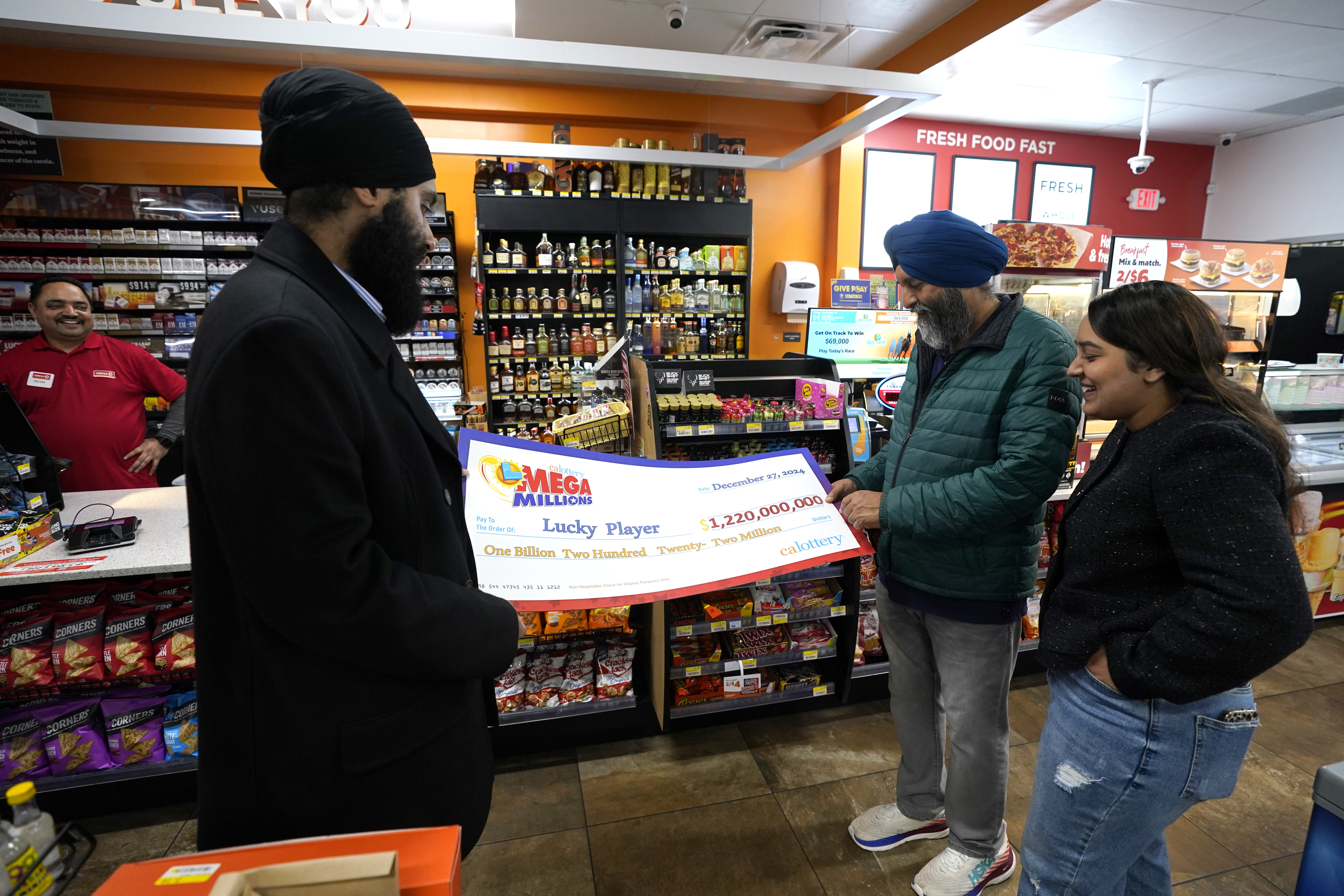 Ishar Gill, left, his father Jaspal Singh, center and his sister Jaspeet Gill right, look over the enlarged image of a check, Saturday, Dec. 28, 2024, that will be presented to the winner of $1.22 billion Mega Millions Lotto ticket that was purchased at their family's store in Cottonwood, Calif., Saturday, Dec. 28, 2024. (AP Photo/Rich Pedroncelli).