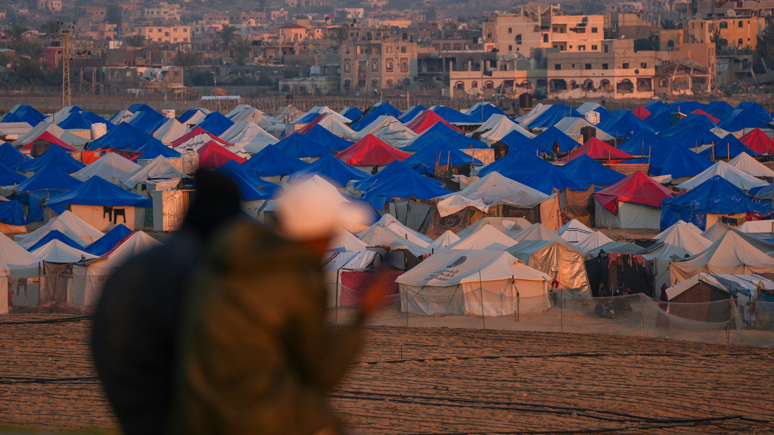 Two people sit at sunset overlooking a tent camp for displaced Palestinians in the outskirts in the central Gaza Strip town of Khan Younis Wednesday, Jan. 1, 2025. (AP Photo/Abdel Kareem Hana)