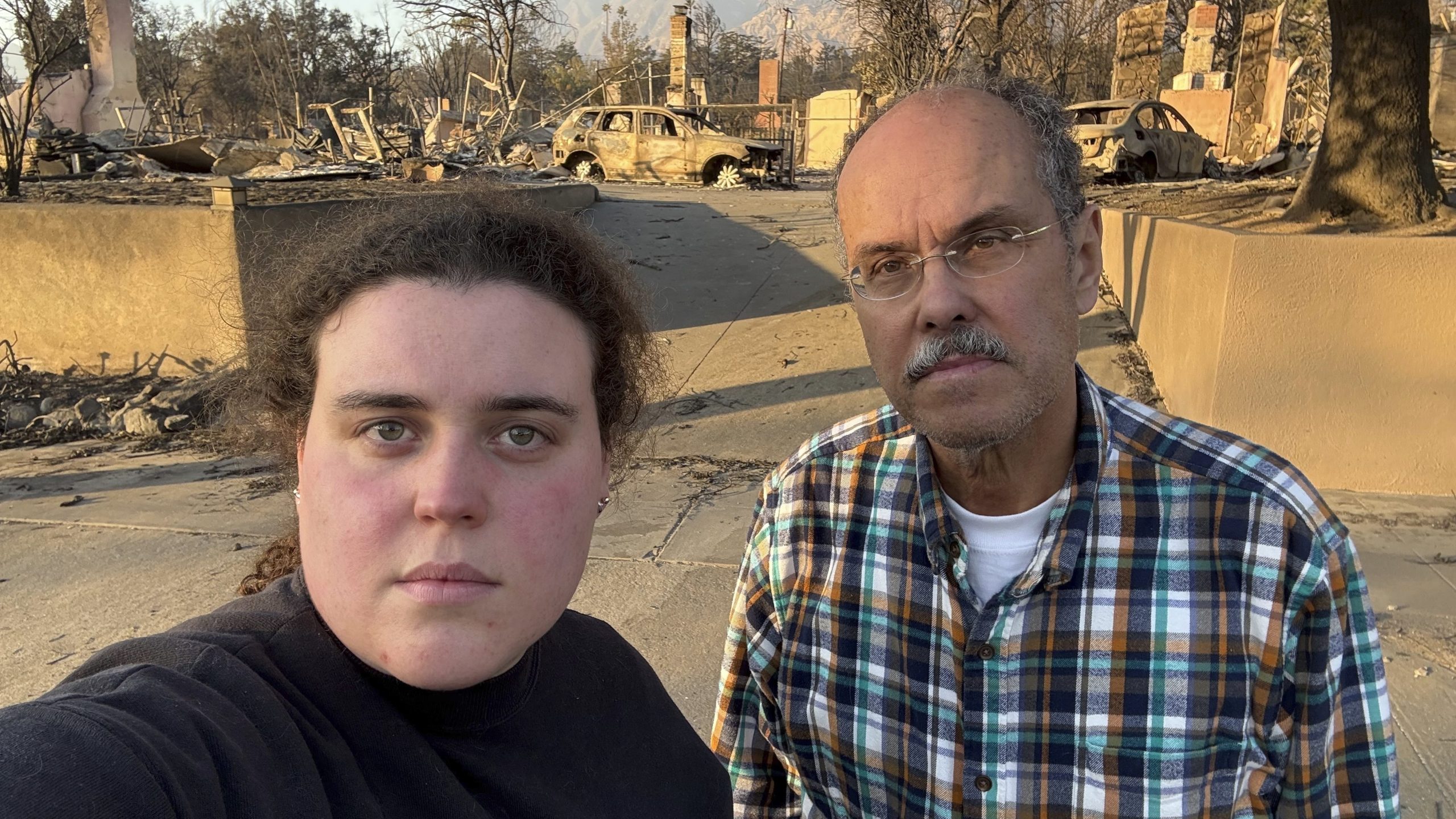 Vanessa Prata and her father, Aluizio Prata, pose for a self-portrait in Altadena, Calif., on Saturday, Jan. 11, 2025, with damage from the Eaton fire behind them. (Vanessa Prata via AP)