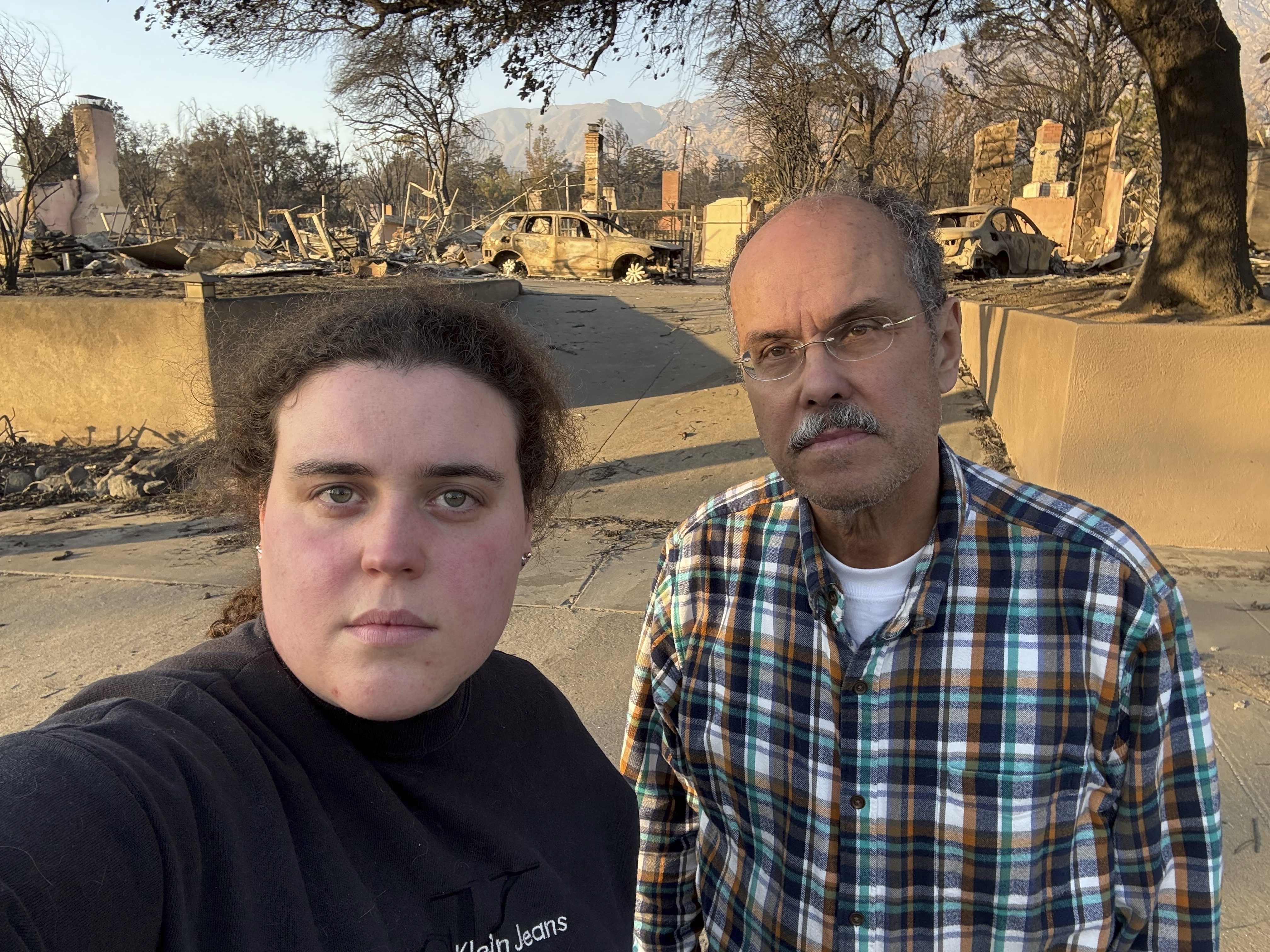 Vanessa Prata and her father, Aluizio Prata, pose for a self-portrait in Altadena, Calif., on Saturday, Jan. 11, 2025, with damage from the Eaton fire behind them. (Vanessa Prata via AP)